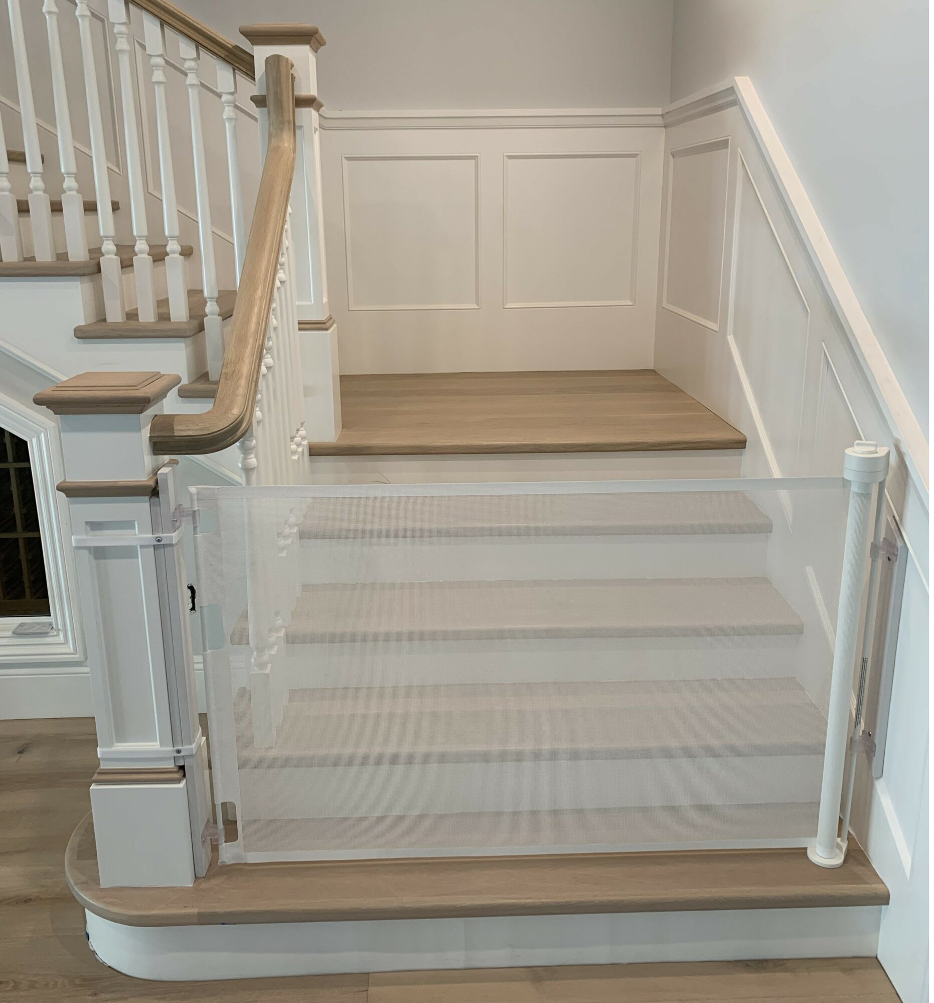 Staircase with wooden handrail and glass panels, leading to a landing with wood flooring, white wall paneling, and a staircase to the right.