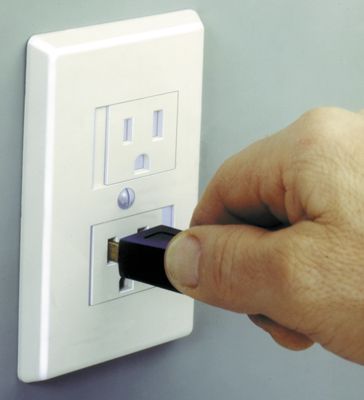 Hand plugging an Ethernet cable into a wall outlet with a power socket and USB port, on a light-colored wall.
