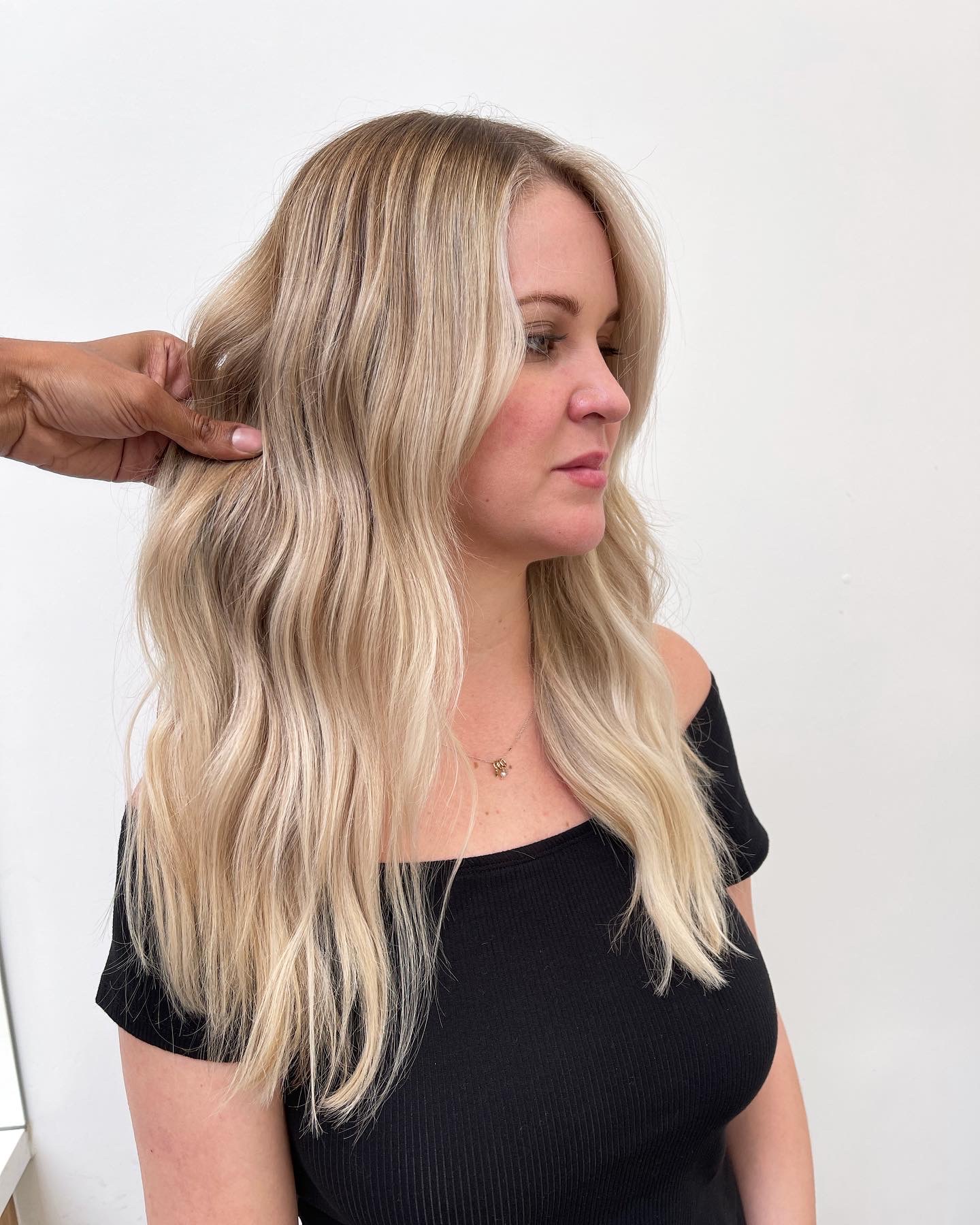 Woman with long wavy blonde hair wearing a black top, looking to the side, against a plain background.