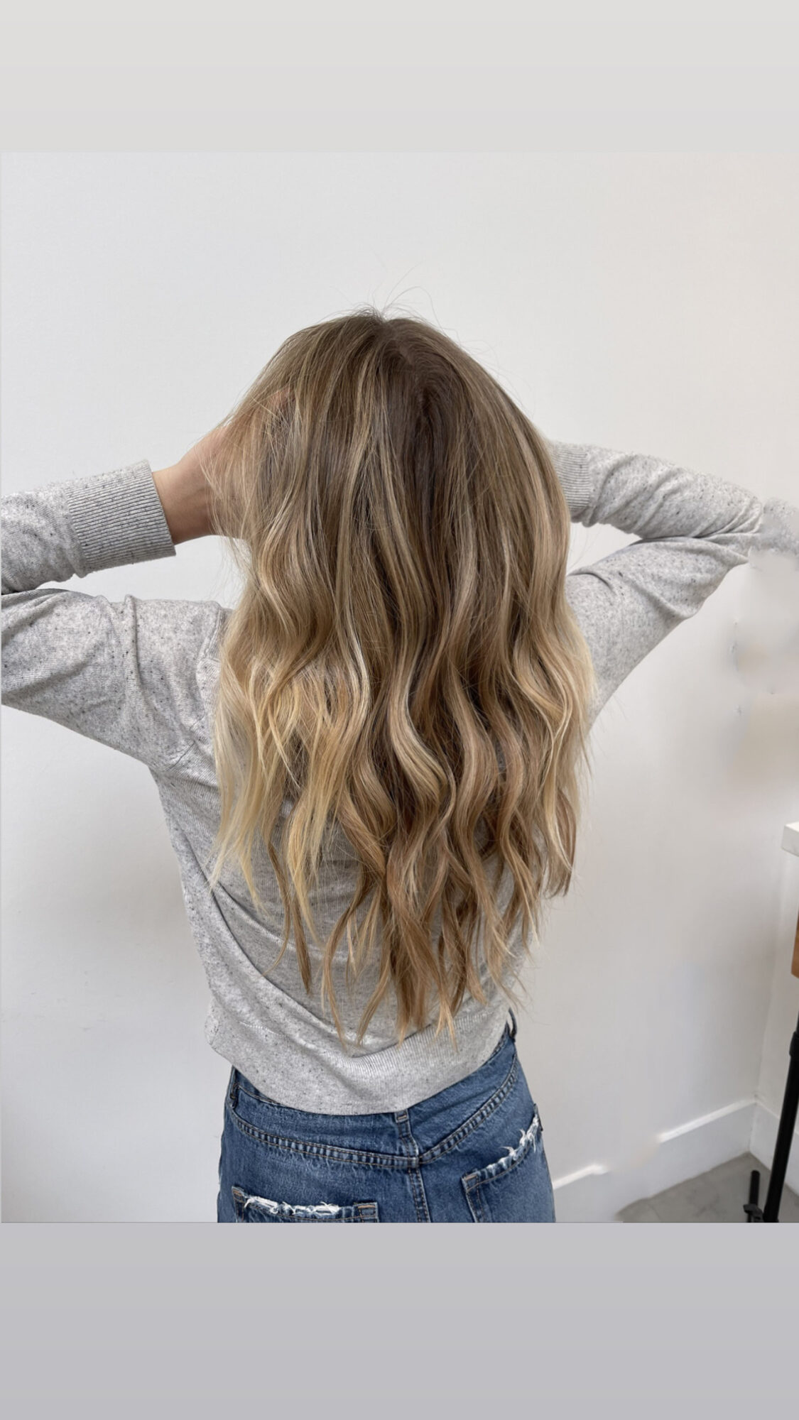 Person with long, wavy blonde hair, wearing a gray top and jeans, facing away from the camera, hands behind head.