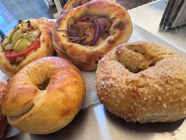 Four bagels with various toppings and coatings on a tray, some with fruit or sweet toppings, one plain, and one with a sugar coating.
