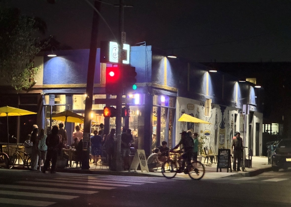 Night scene of a busy street corner with people, bicycles, and a building with illuminated signs and yellow umbrellas.