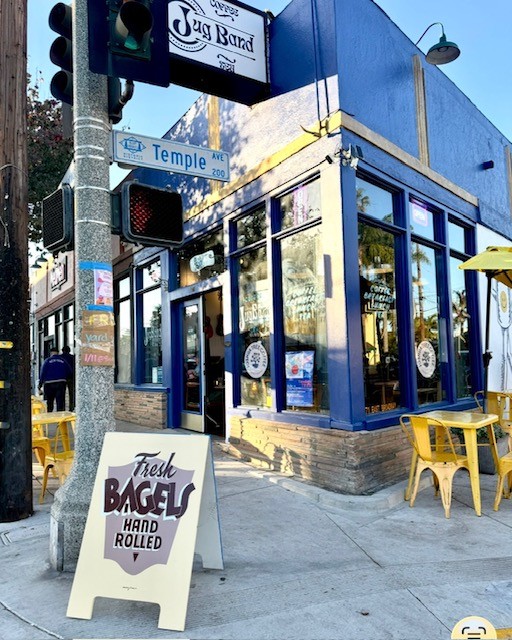 Blue building on corner with yellow chairs outside, street sign reads 'Temple', sandwich board advertises bagels, clear sky, urban setting.