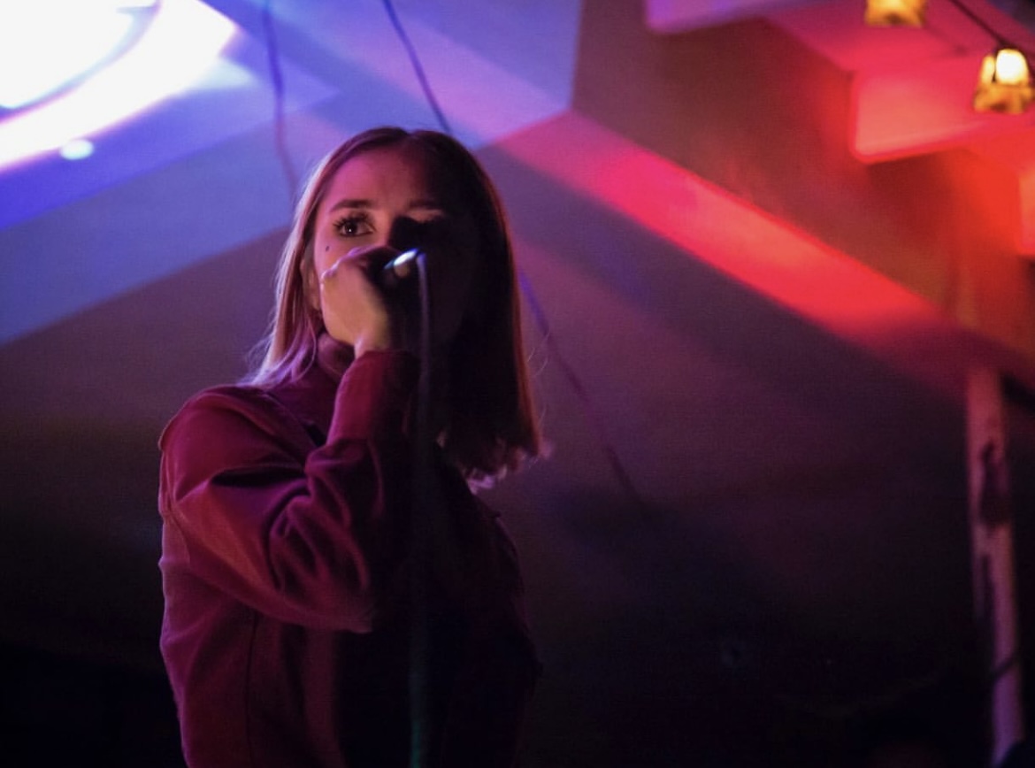 Young woman with shoulder-length hair holding a microphone, illuminated by colorful stage lights, in a dimly lit venue.