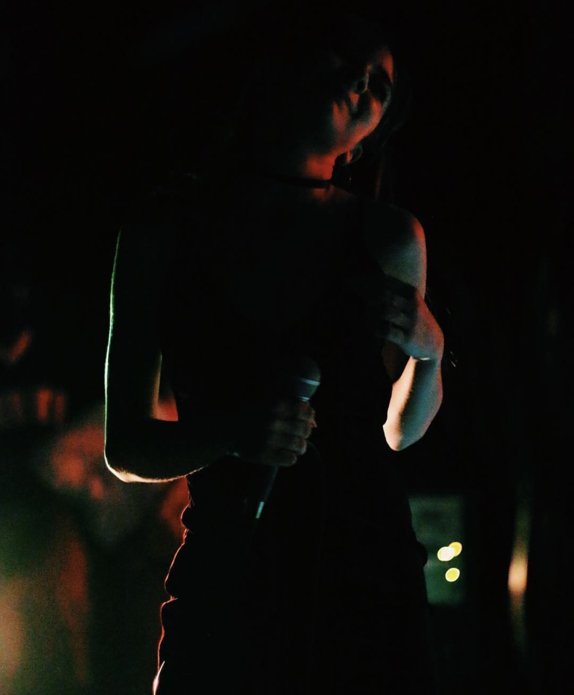 Person holding a microphone in a dark setting with minimal lighting, face partially visible, wearing dark clothing.