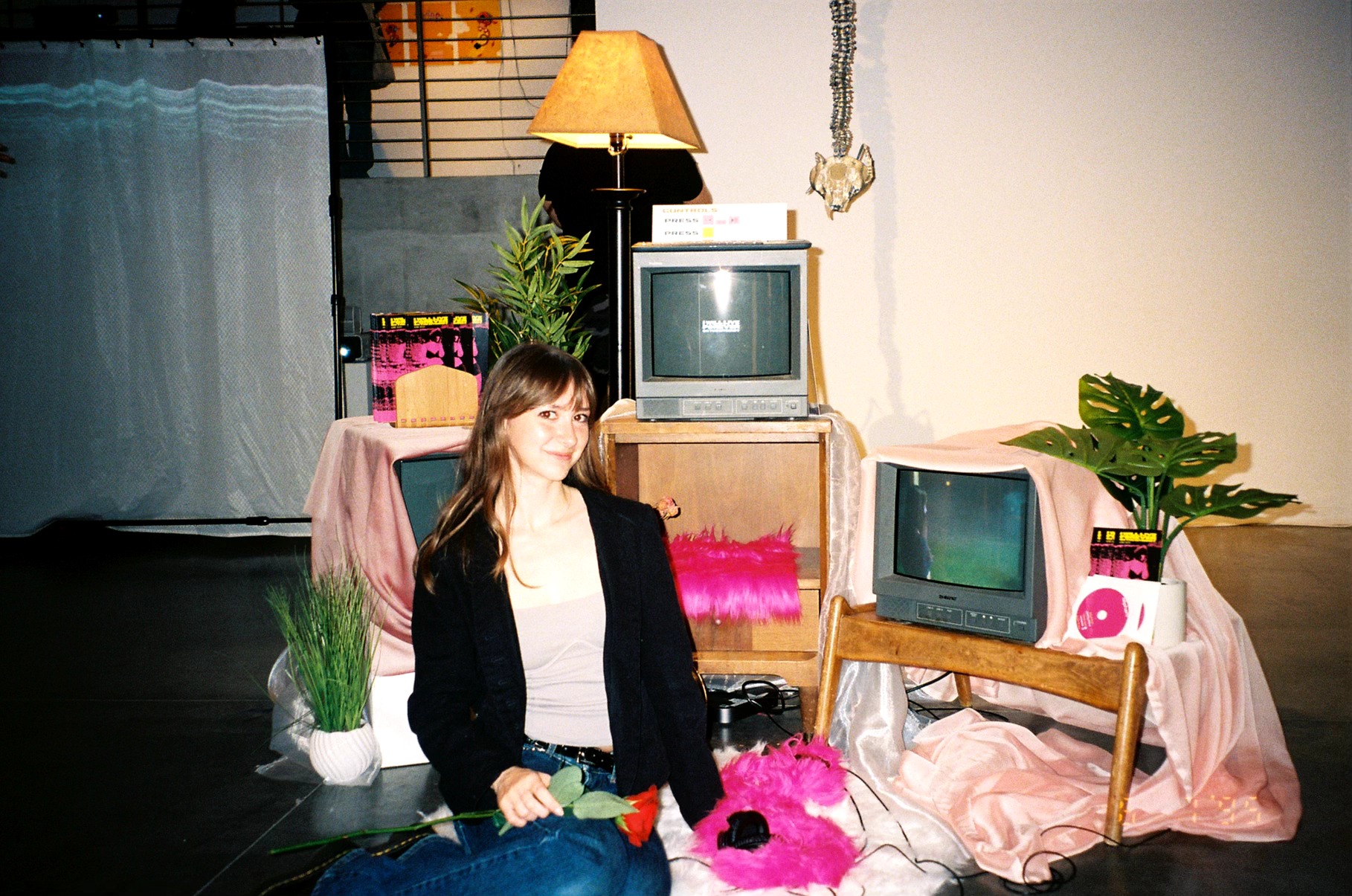 Woman sitting on the floor surrounded by vintage TVs, plants, and pink decorations in a cozy room.