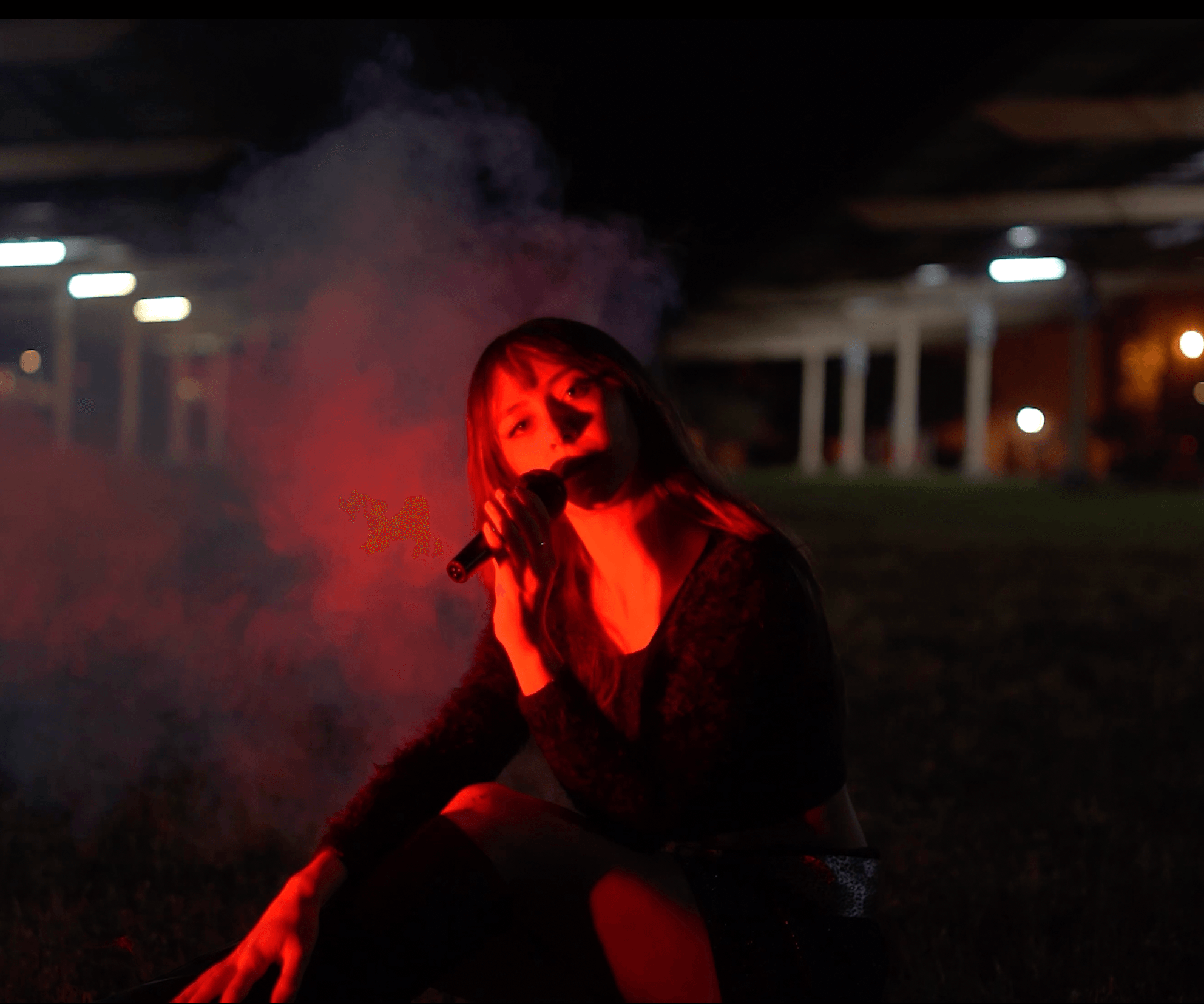 Person with long hair sitting outdoors at night, illuminated by red and orange light, with smoke nearby and a dark background.