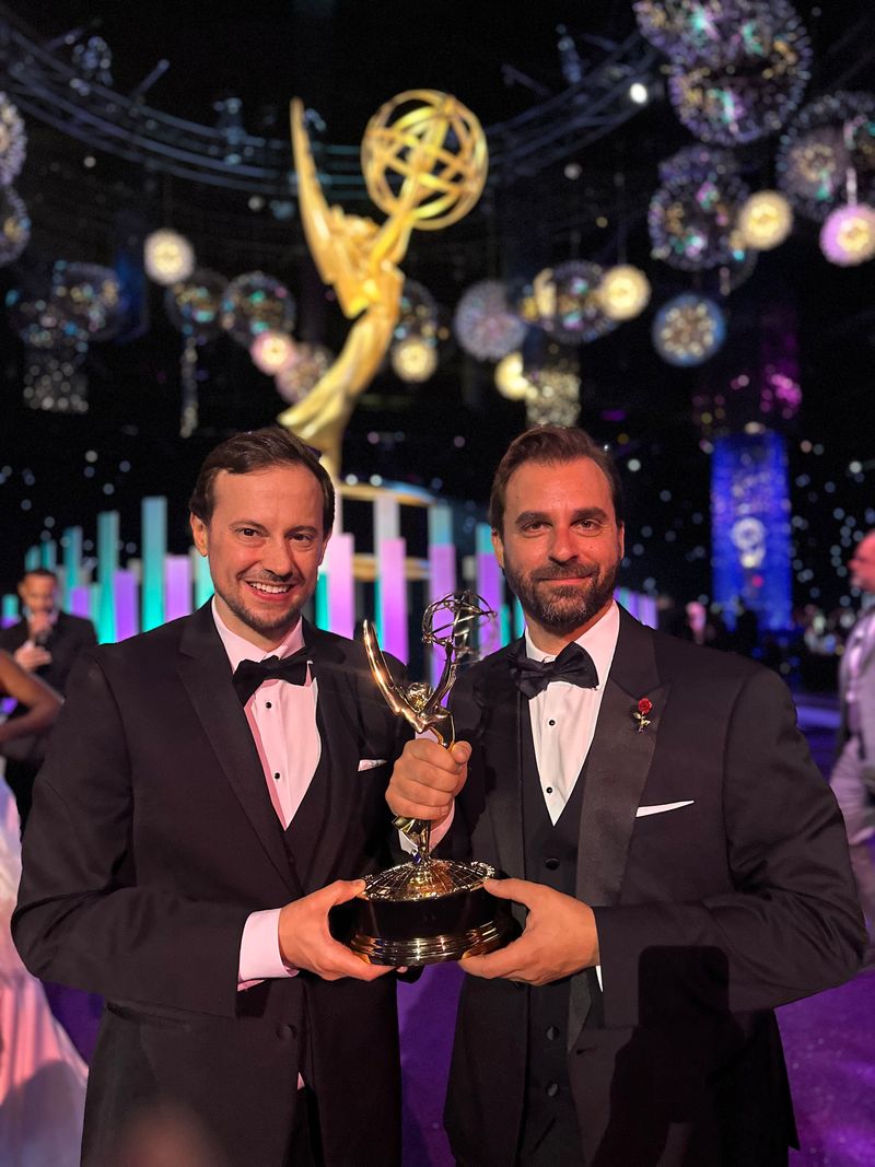 Two men in tuxedos holding an Emmy award on stage with a large Emmy statue and colorful decorations in the background.