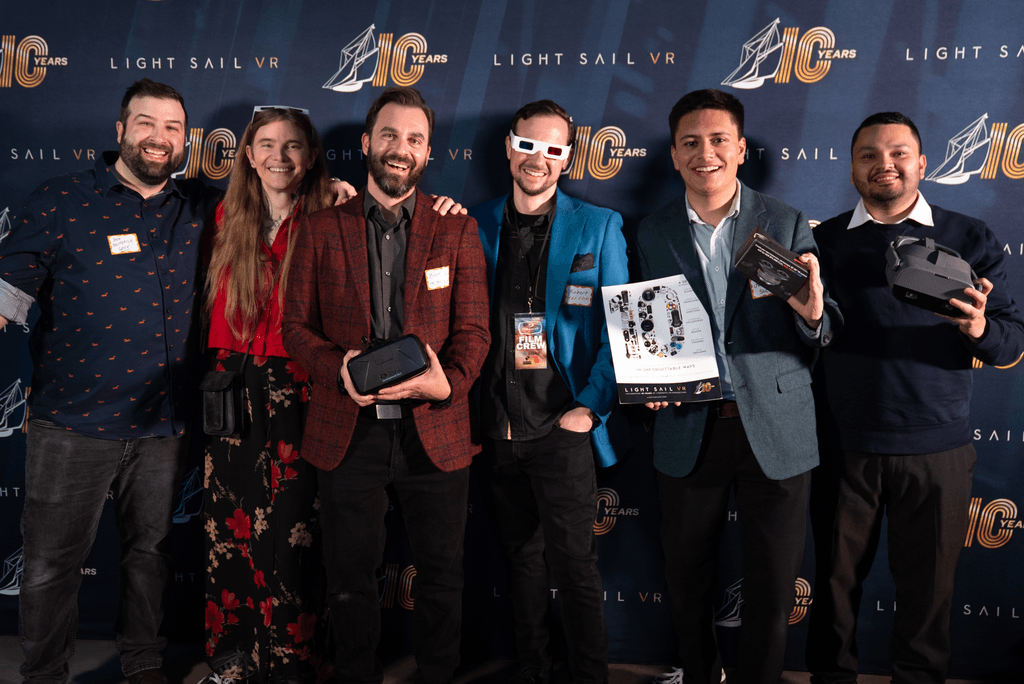 Group of six people smiling at an event, holding virtual reality headsets and boxes, with a blue backdrop featuring logos.