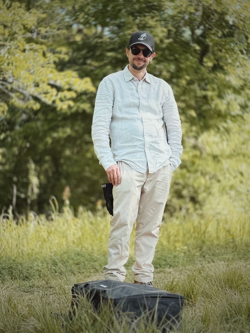 Man standing outdoors on grass with trees in background, wearing sunglasses, a cap, light-colored shirt and pants, smiling.