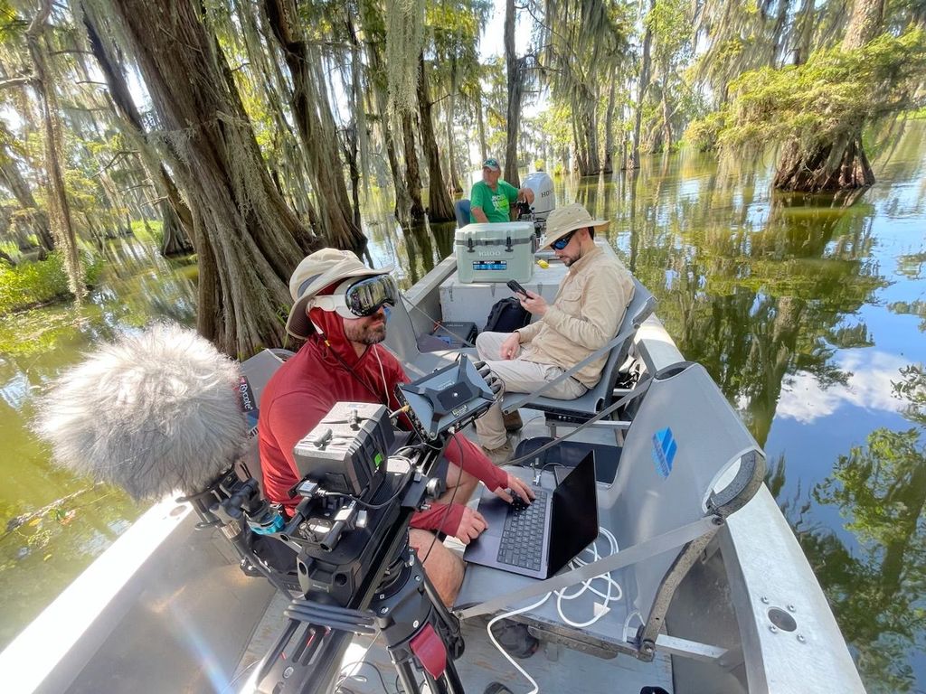 Scientists in outdoor gear working on equipment on a boat in a swampy area with trees and water.