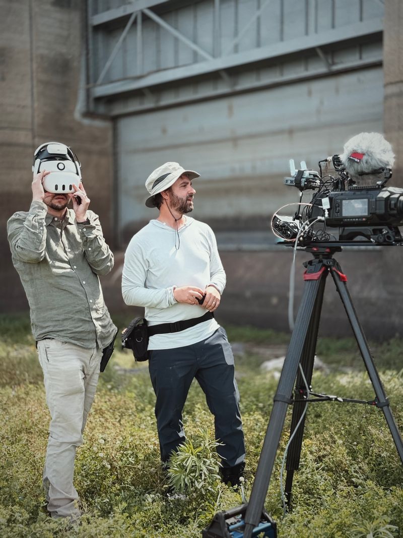 Two men stand outdoors near a camera on a tripod, one wearing a virtual reality headset, the other looking at the camera, with a bridge in the background.