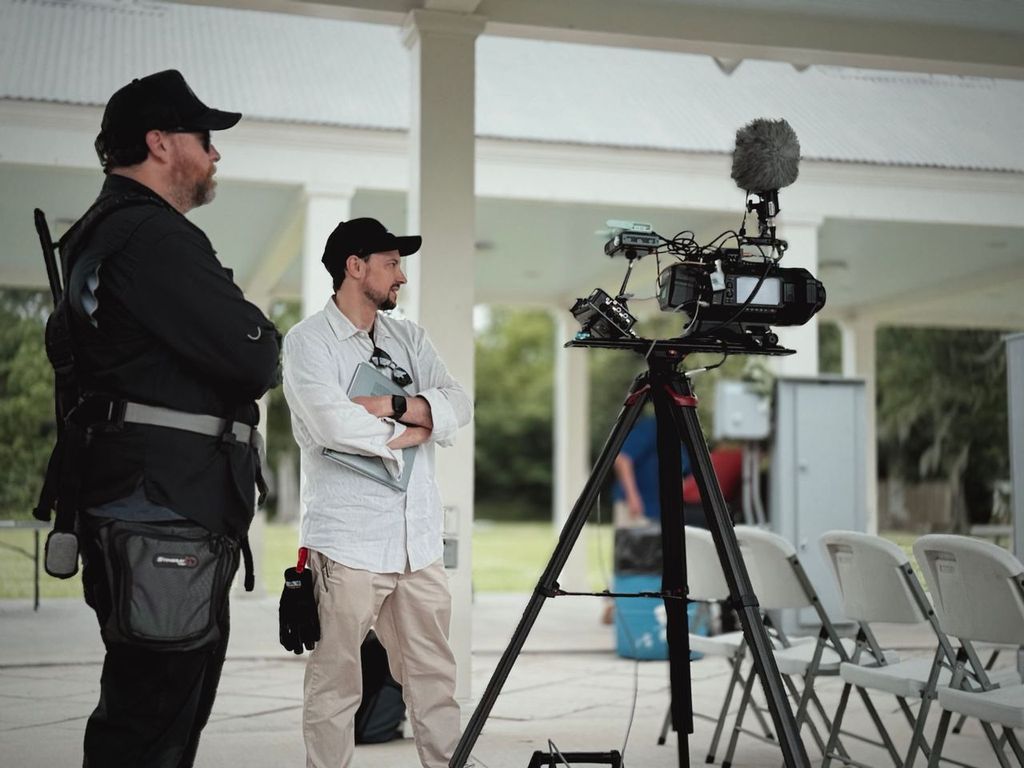Two men standing outdoors near a camera on a tripod, with chairs and a building in the background.