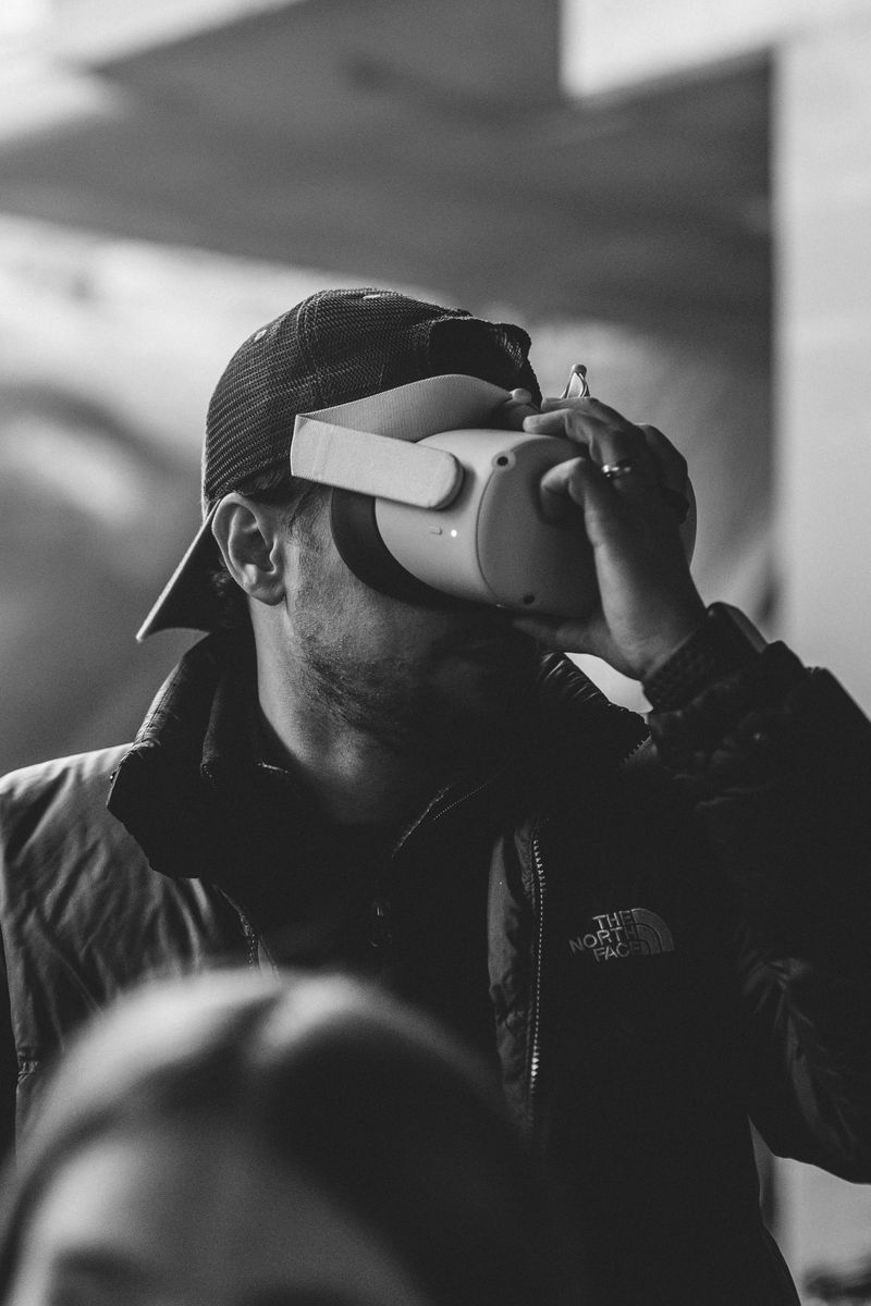 Person wearing a cap and virtual reality headset, holding the device with one hand, in black and white.