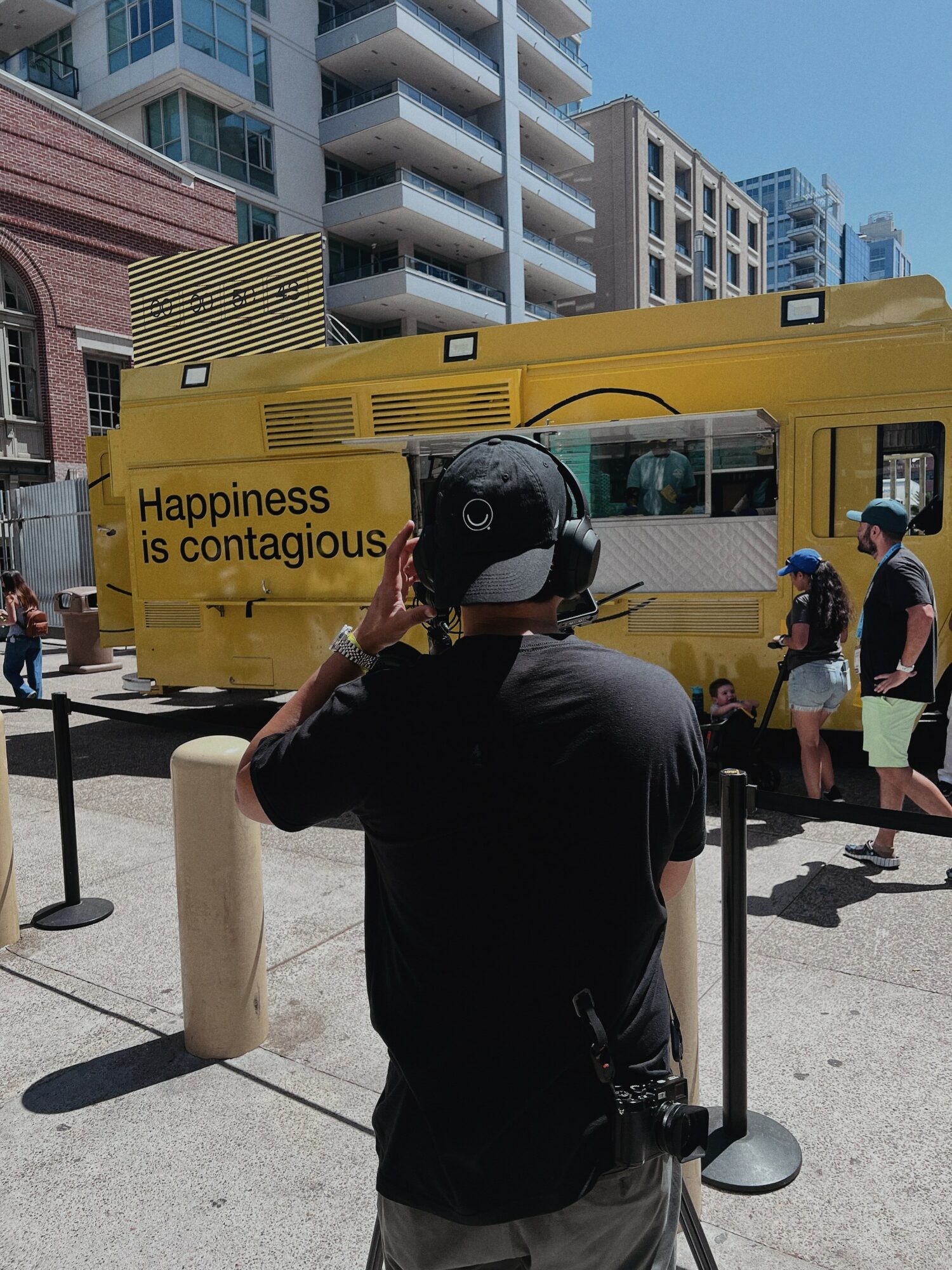 Person wearing a black shirt and cap talking on a phone in front of a yellow bus, city buildings in background.