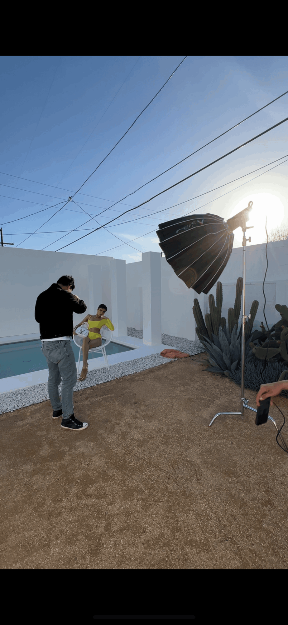 Person taking photo near pool with a large umbrella and cactus, bright sunlight, outdoor setting.