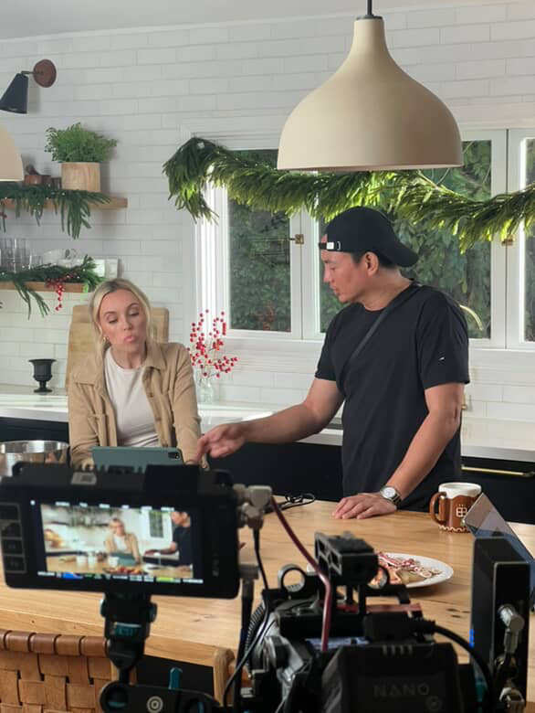 Two people in a kitchen, one standing and one sitting, with a camera recording them, and a large hanging light above.