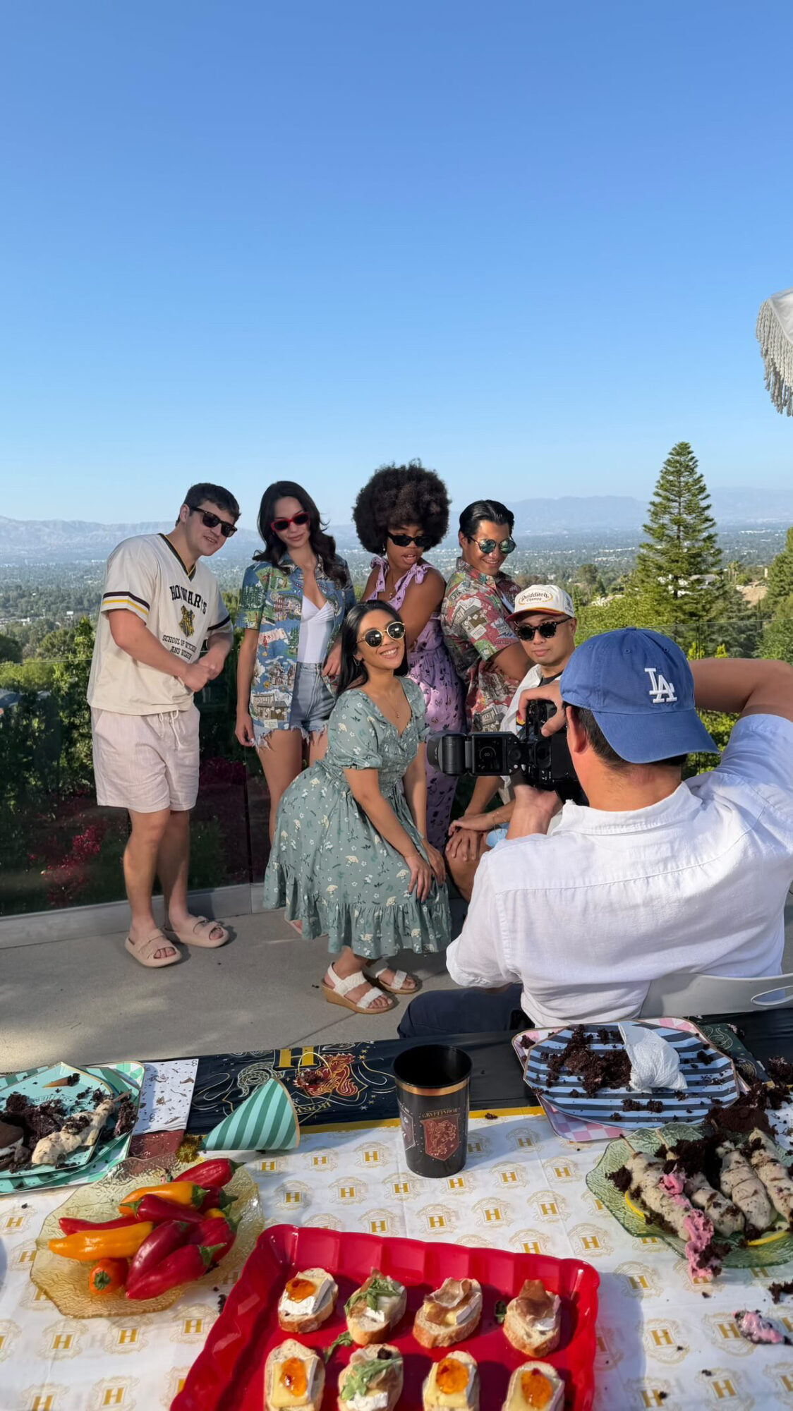 Group of six people outdoors, some standing and one sitting, with a table of food in front, under a clear blue sky.