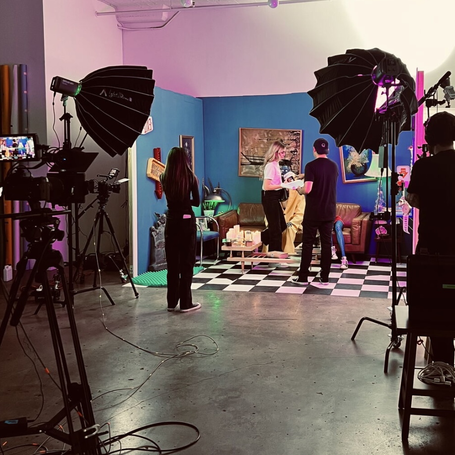 People in a studio with colorful walls, furniture, and lighting equipment, filming or photographing a scene with a person on set.