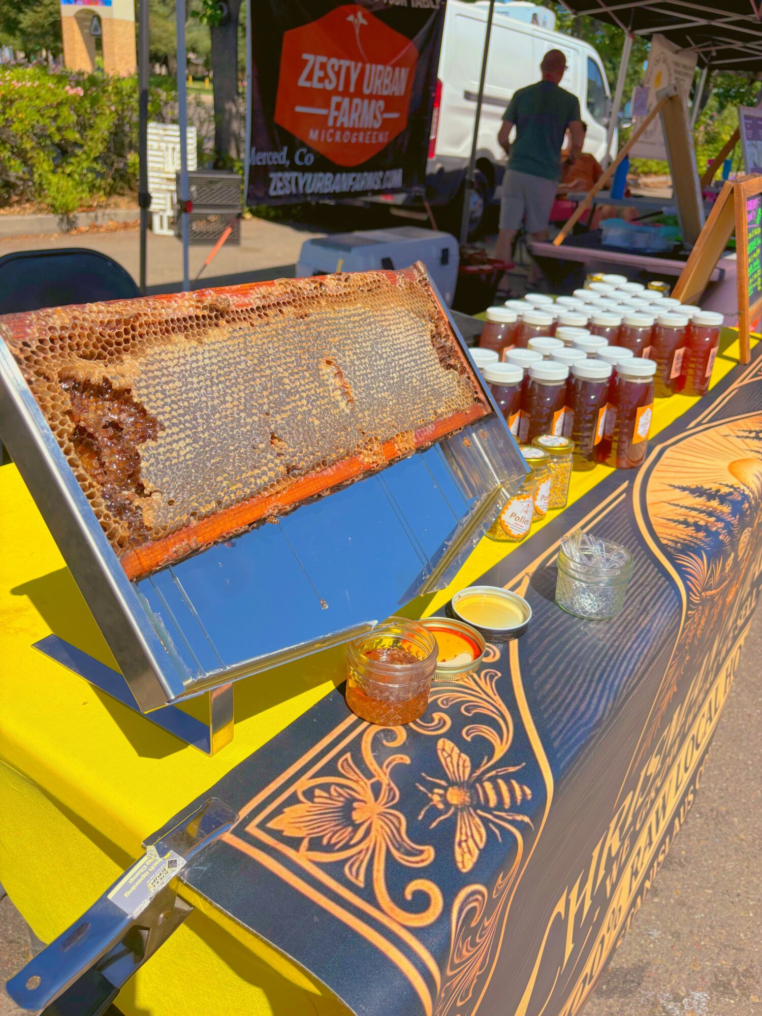 Honeycomb frame on a table with honey in a jar, at an outdoor market stall with drinks and people in background.