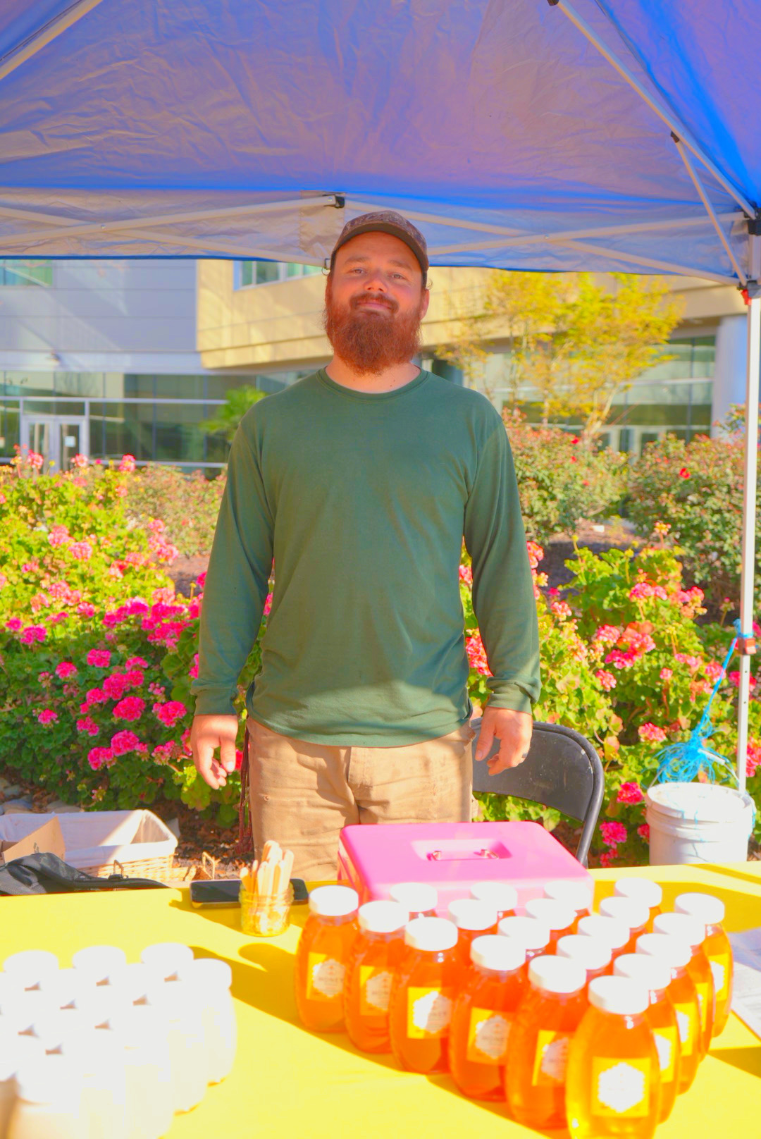Man with a beard and green shirt standing outdoors under a blue canopy, surrounded by pink flowers, with a table of bottled drinks in front.