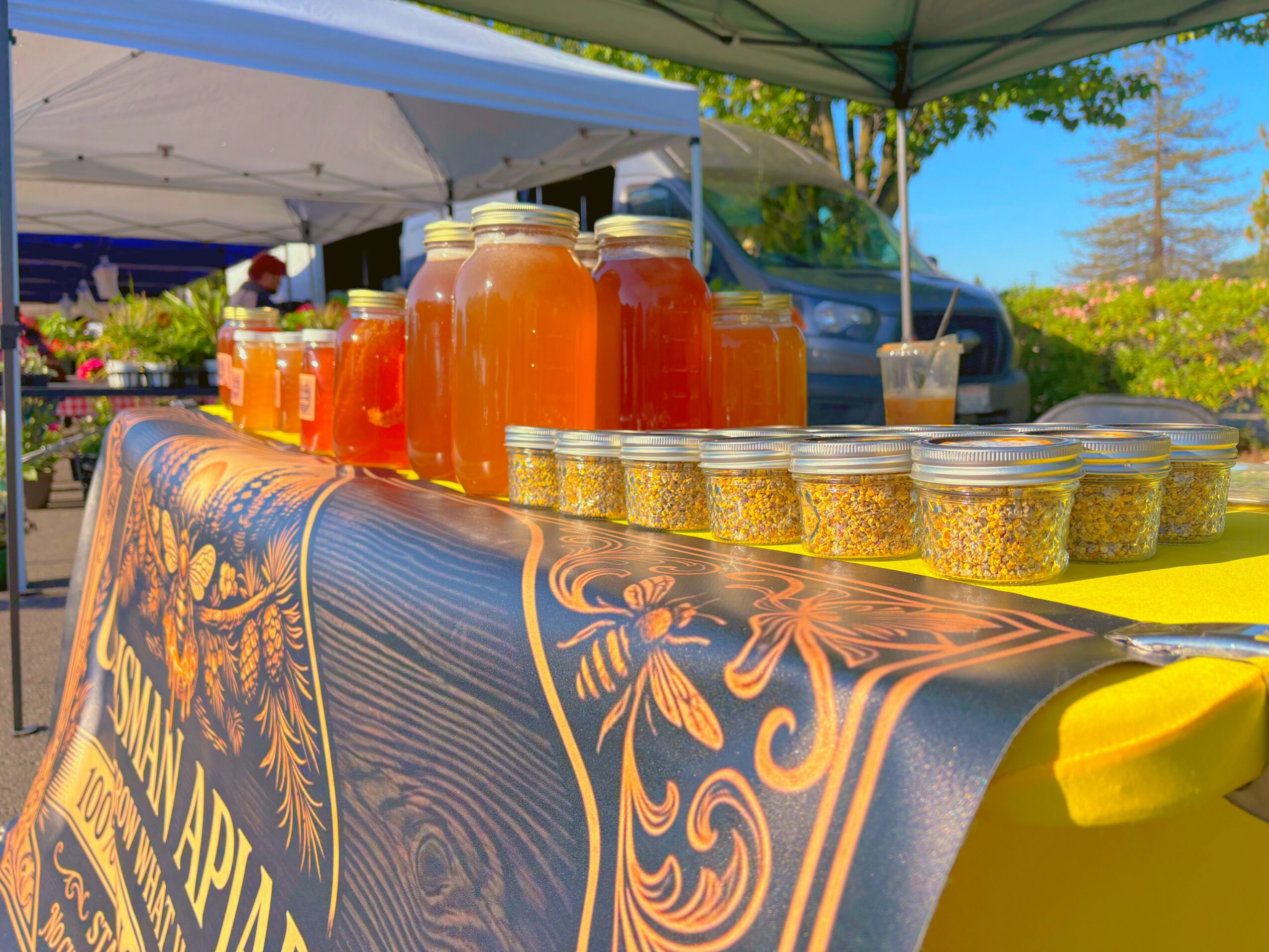 Bottles of orange juice and small containers on a table at an outdoor market stall, with tents and trees in the background.