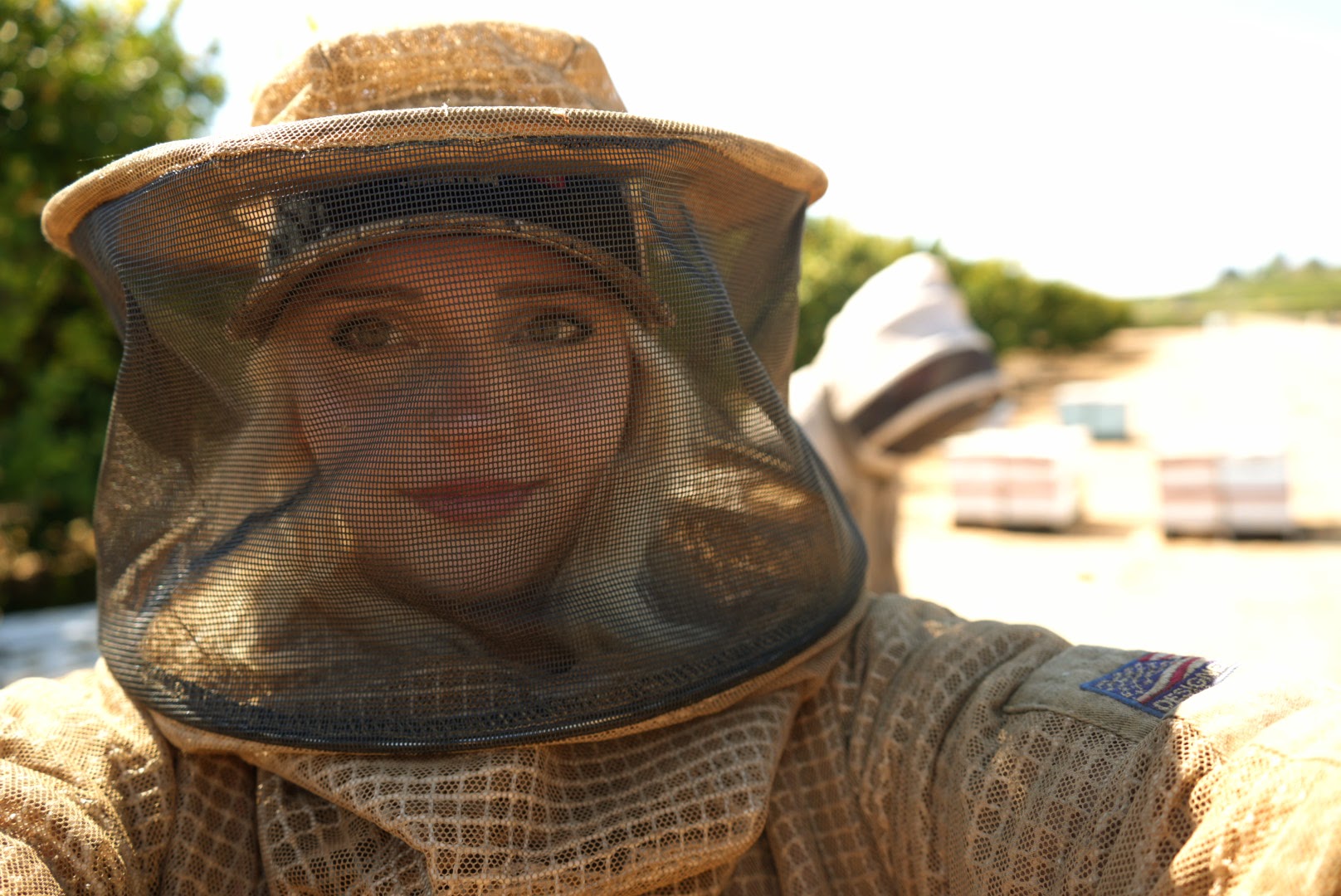 Person wearing a protective beekeeping suit and hat with veil outdoors, smiling, with trees and sky in background.