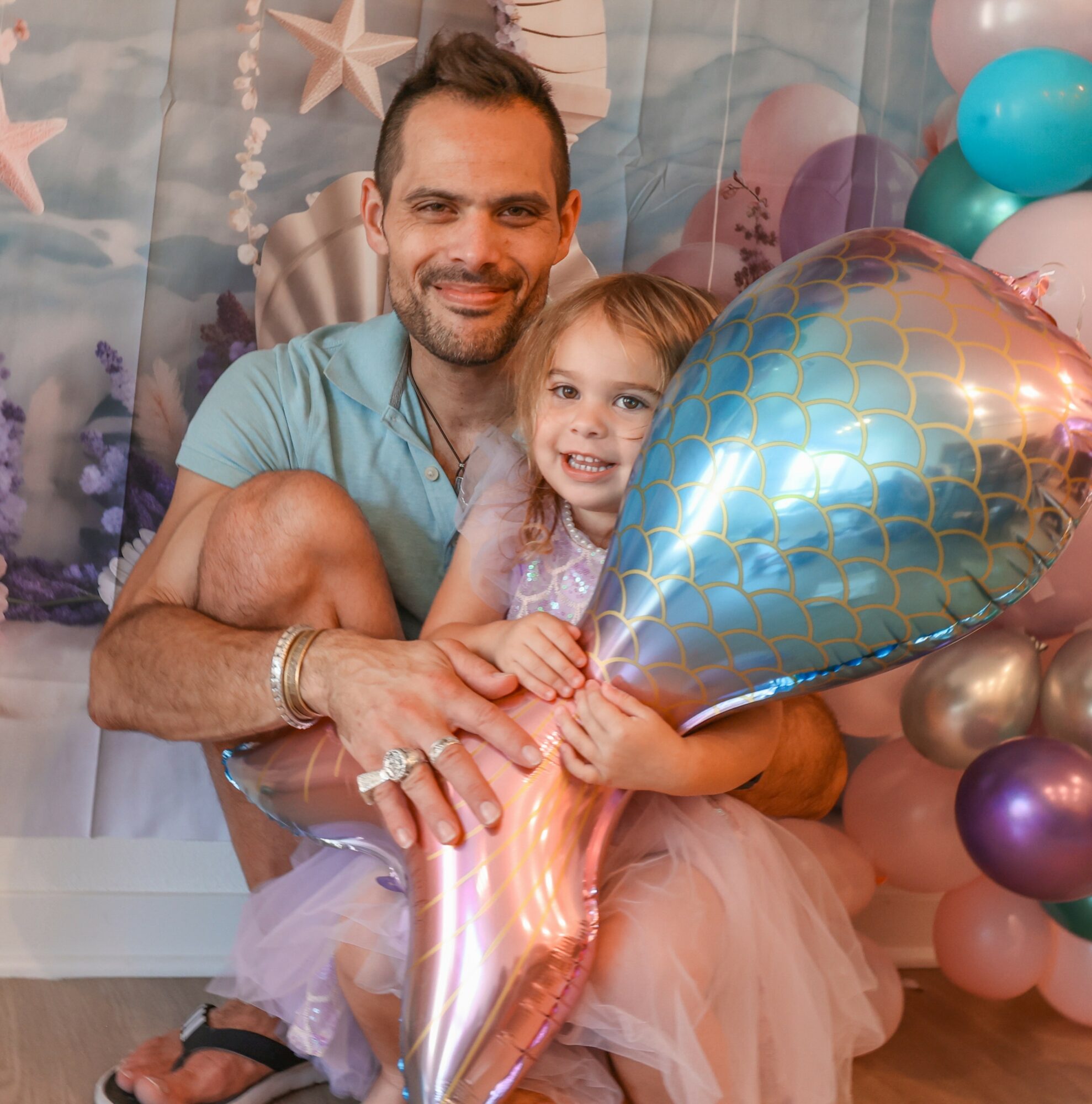 A man and young girl smiling, holding a large shiny balloon, surrounded by colorful balloons and decorations.