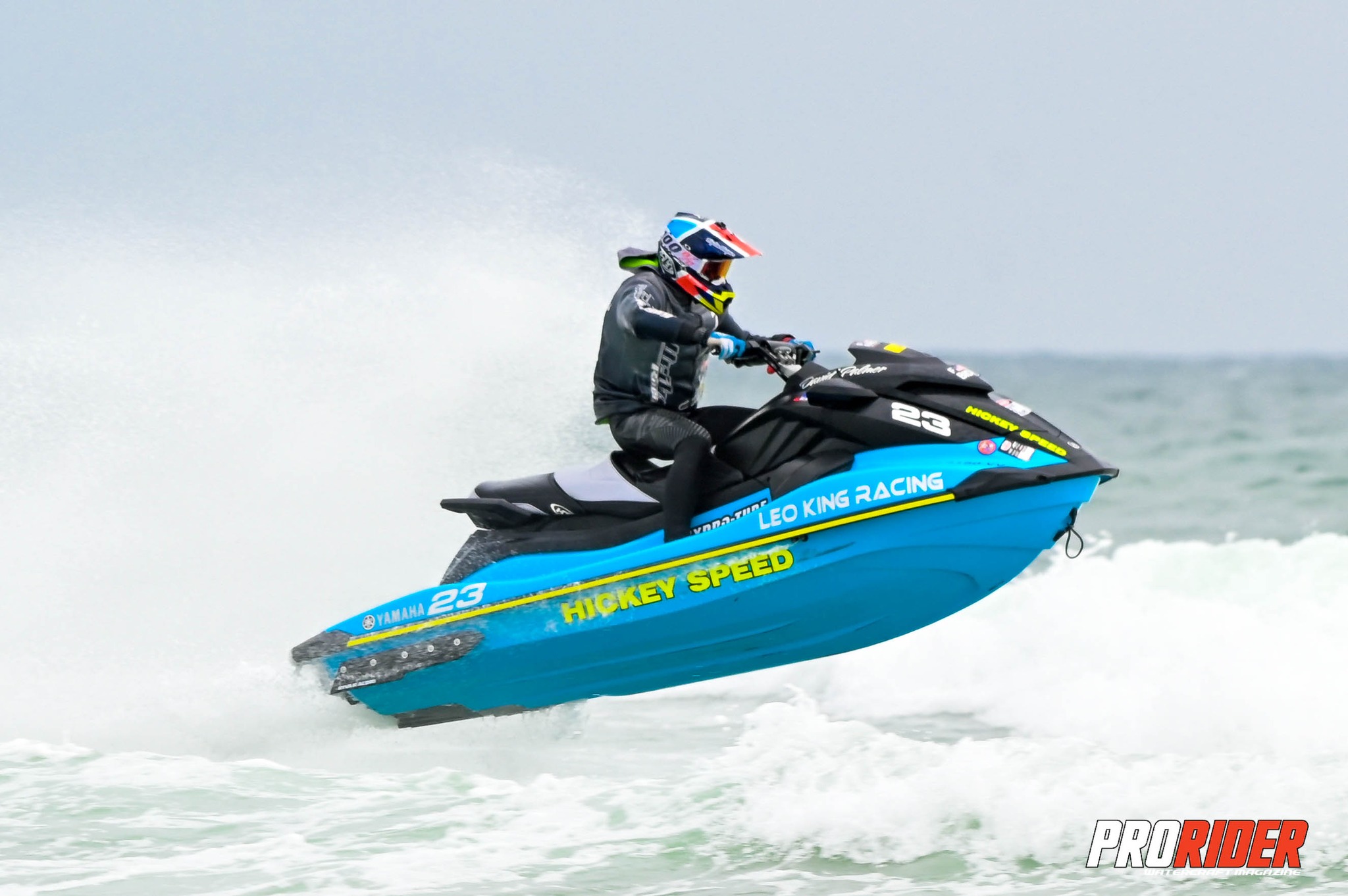 Person riding a blue and black jet ski on water, wearing a helmet and life jacket, with a cloudy sky background.