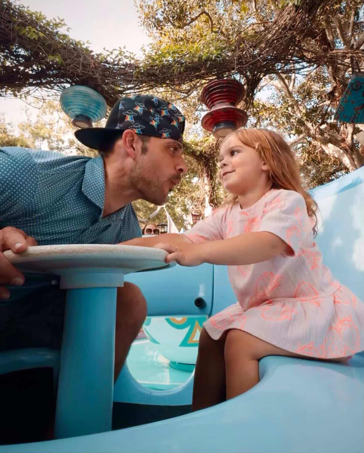 A man and a young girl face each other at a table outdoors, with colorful lanterns hanging above and trees in the background.