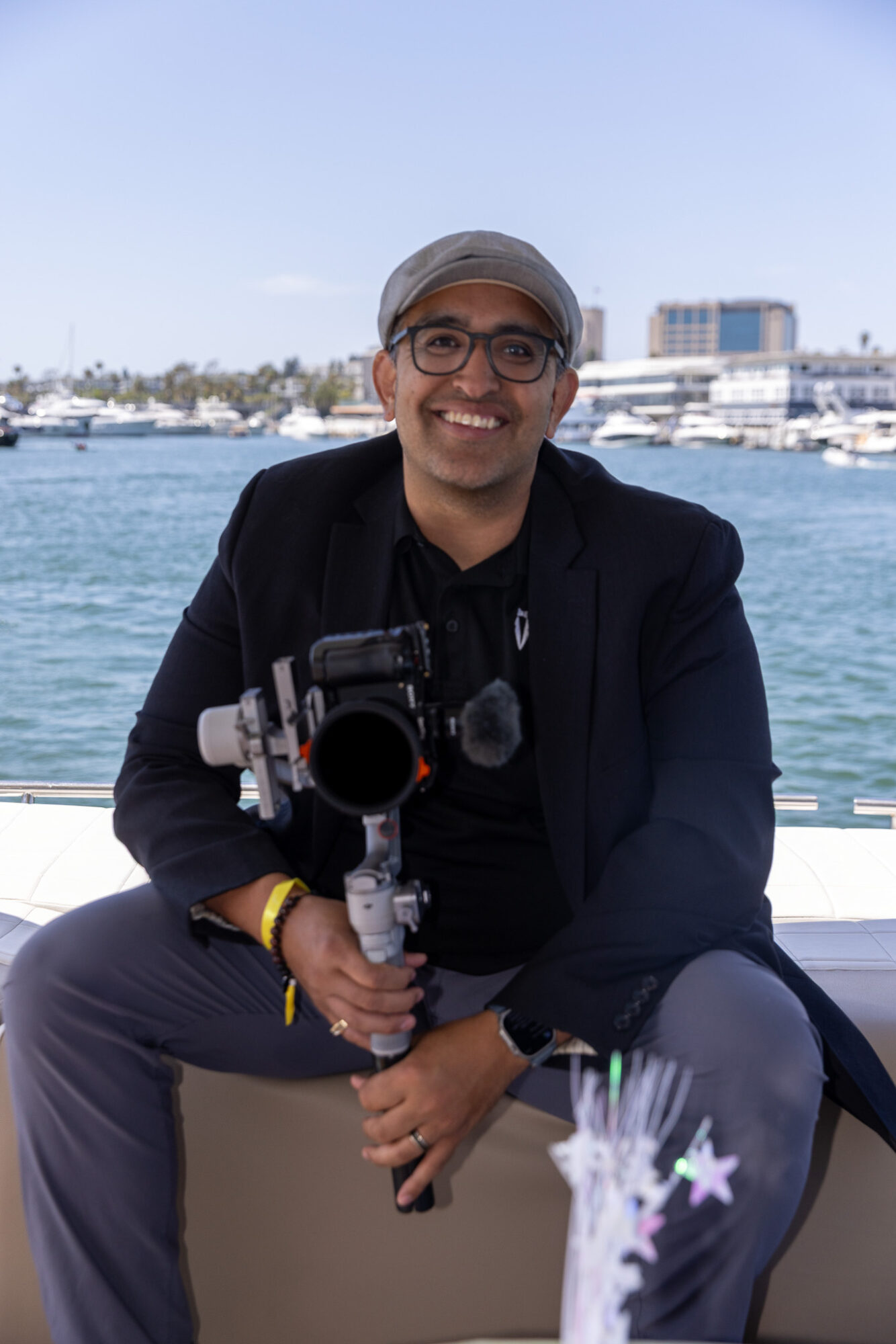 Man smiling with camera equipment, sitting outdoors near water with boats and buildings in background.