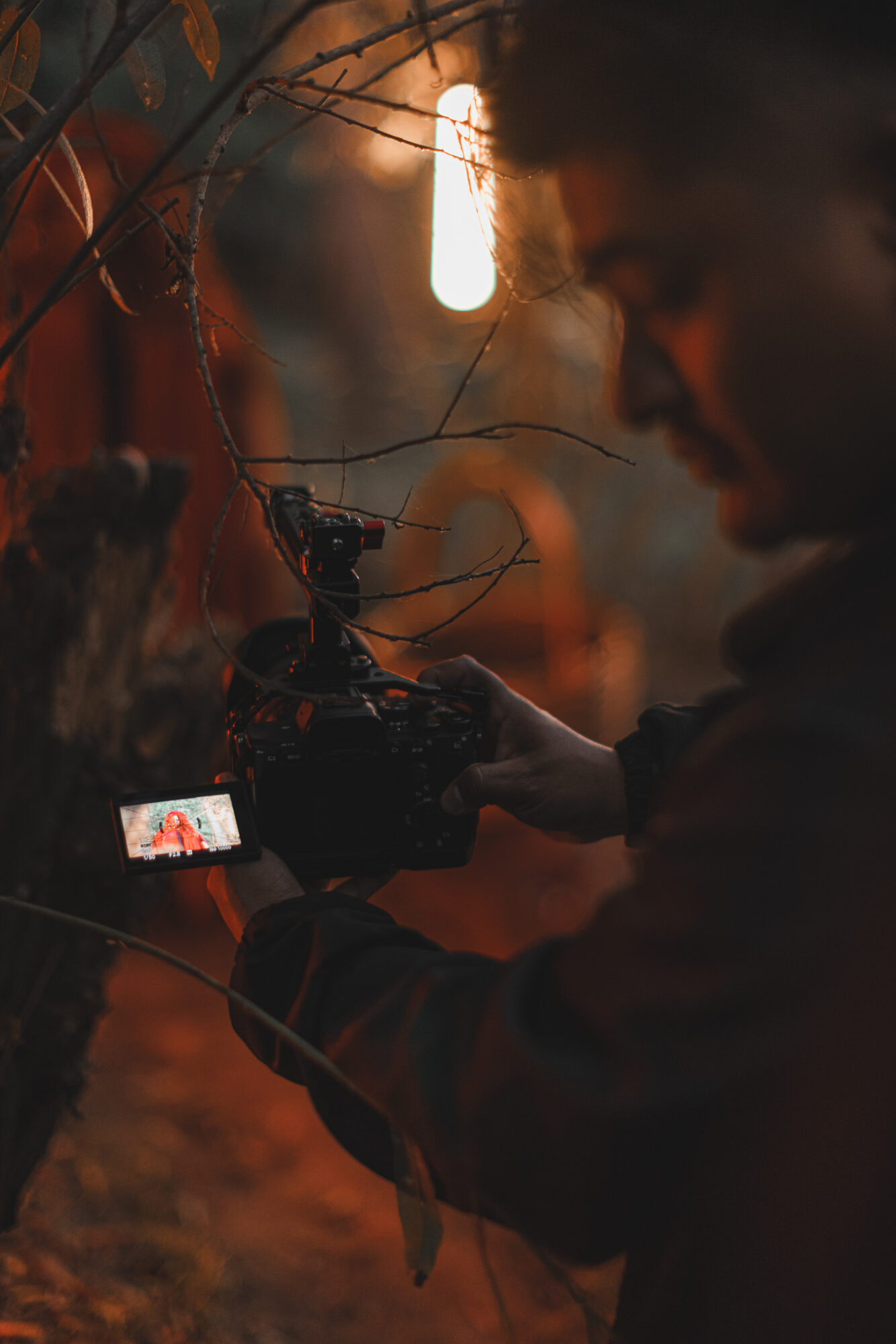 Person holding a camera outdoors at dusk, with a bright light hanging above, and trees in the background.