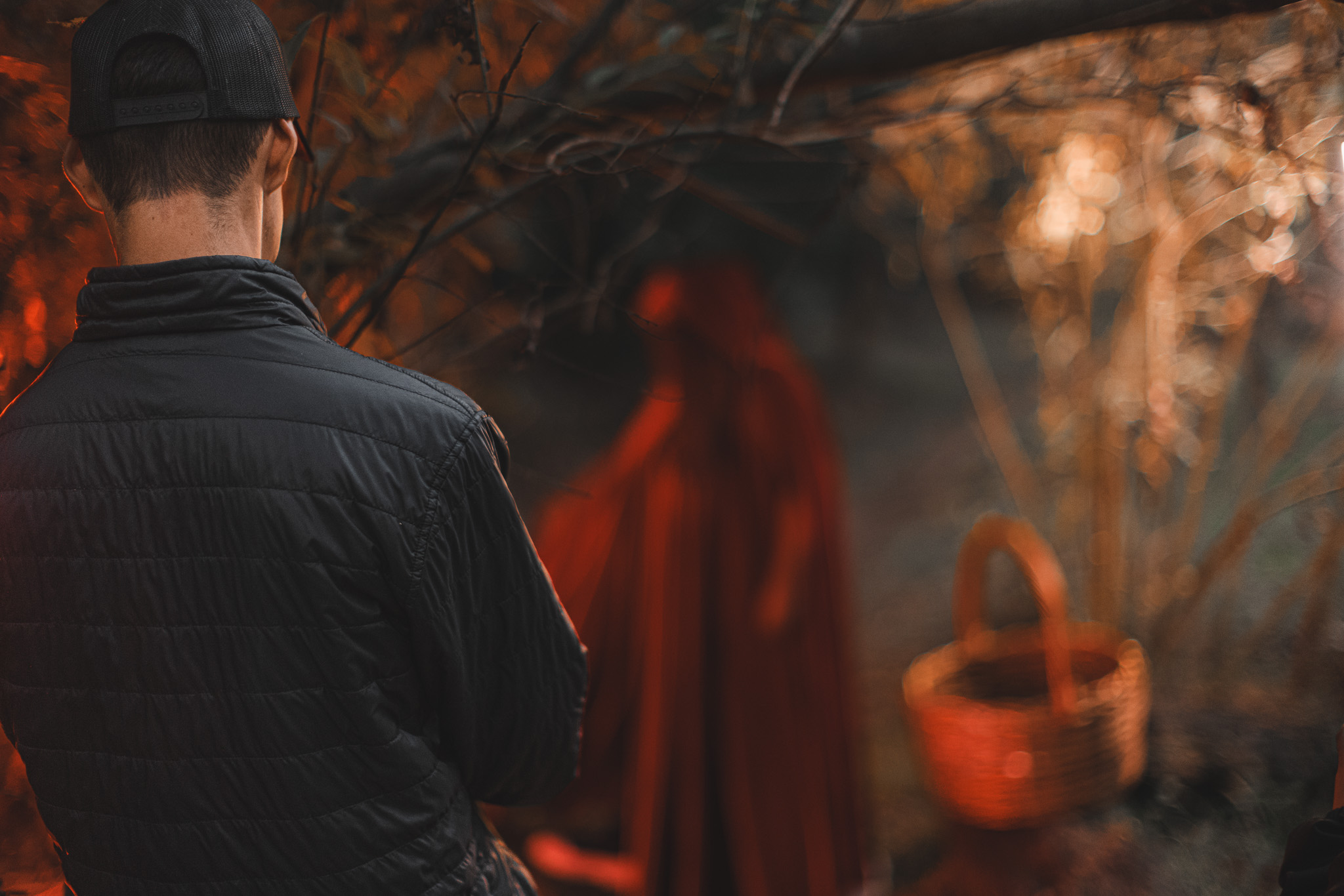 Person wearing a black jacket and cap facing away outdoors with blurred background of trees and a basket.