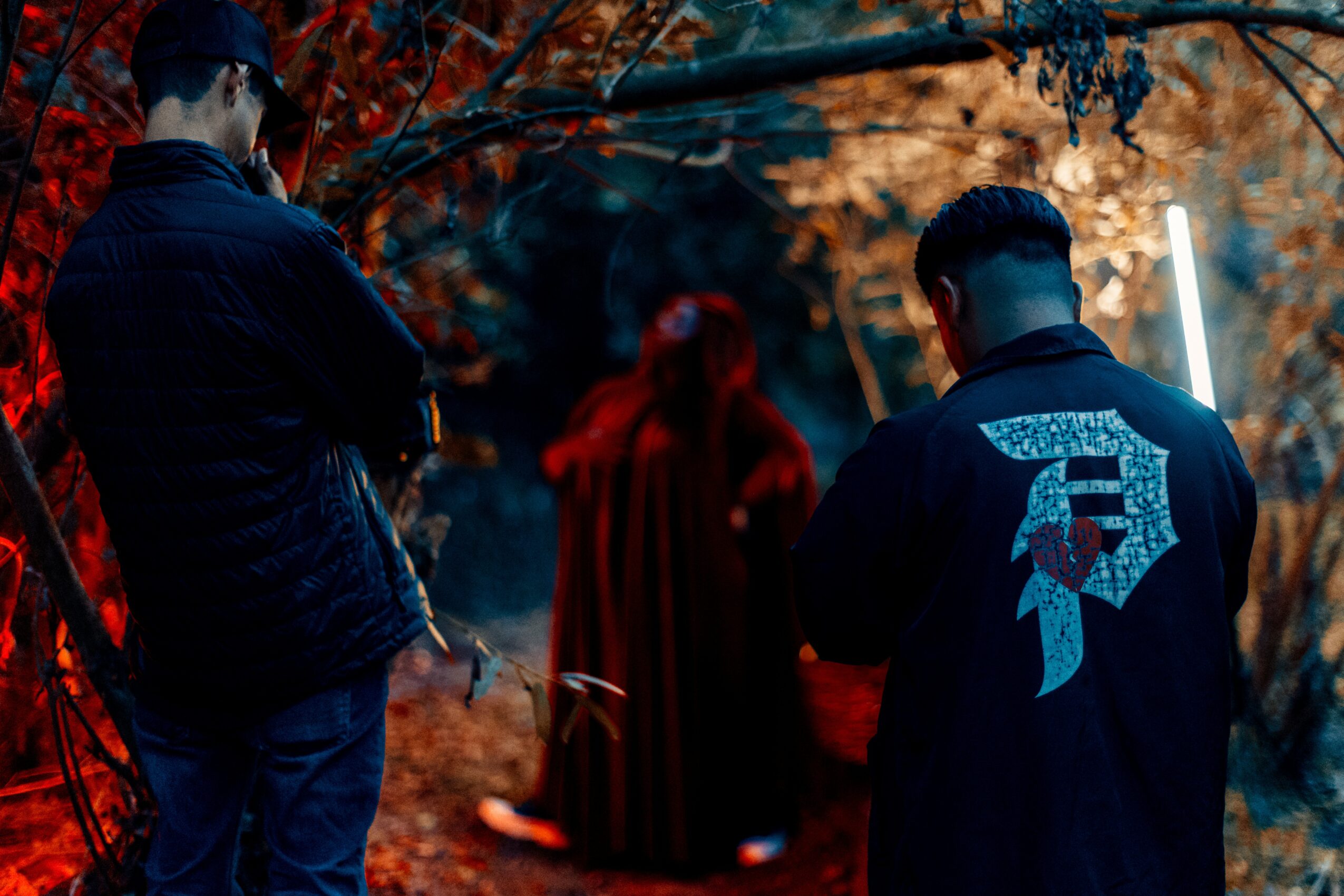 Two people with their backs turned, standing outdoors with autumn trees, observing a person in a red cloak in the background.