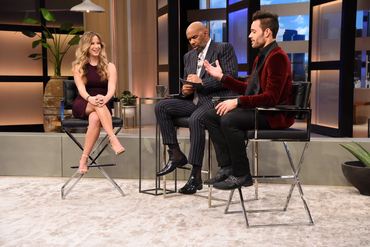 Three people seated on a talk show set with a modern background, two men and a woman smiling and talking.