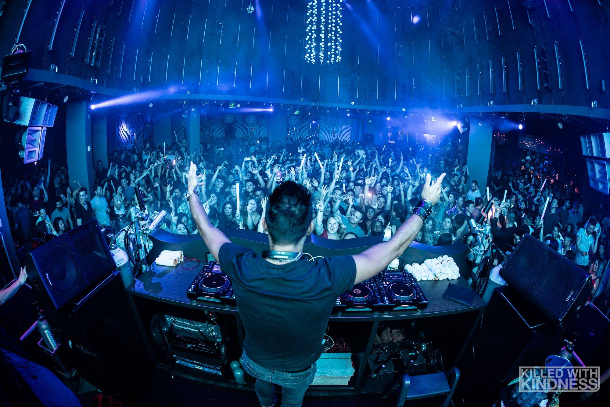 DJ performs at a nightclub with a large crowd dancing and raising hands, blue lighting, and electronic equipment visible.