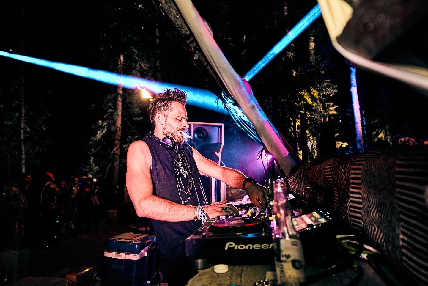 DJ wearing sleeveless shirt and headphones, mixing music at an outdoor night event with colorful lights.