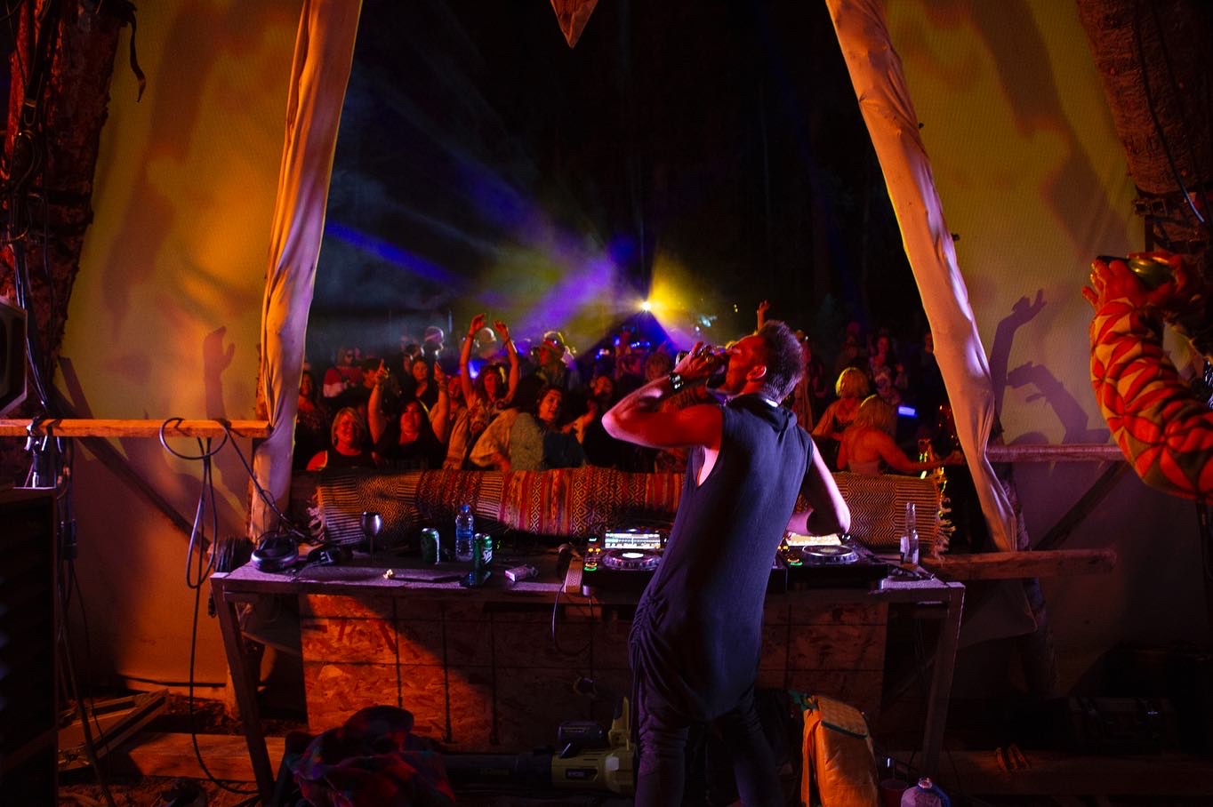 Person DJing at a colorful stage with crowd and vibrant lights, inside a tent-like structure with orange and yellow fabric.