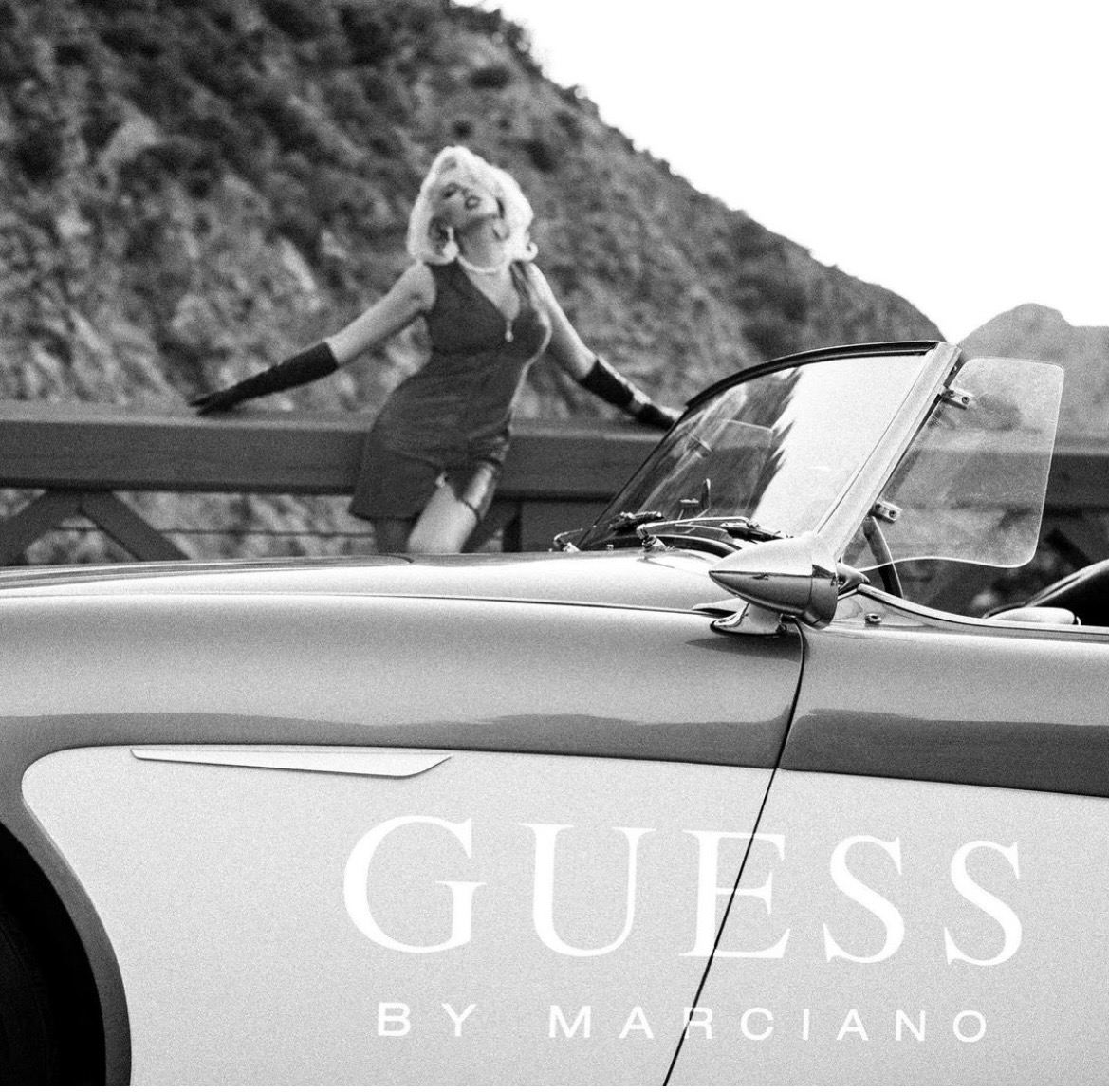 A woman with curly hair and sunglasses leans on a guardrail near a convertible car, with a hillside in the background.