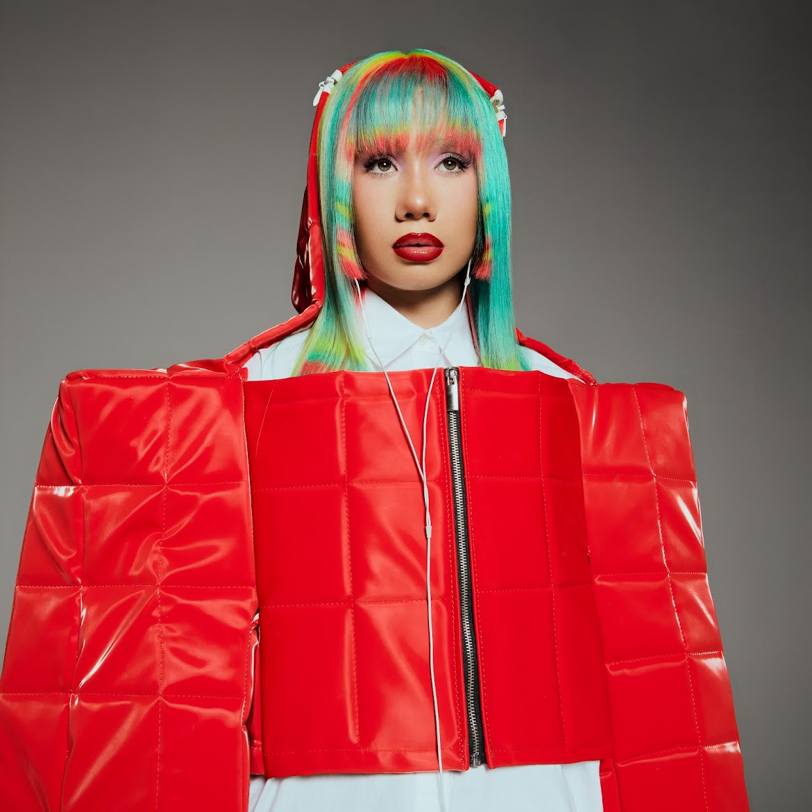 Person with colorful hair wearing a red padded jacket and white shirt, against a gray background.