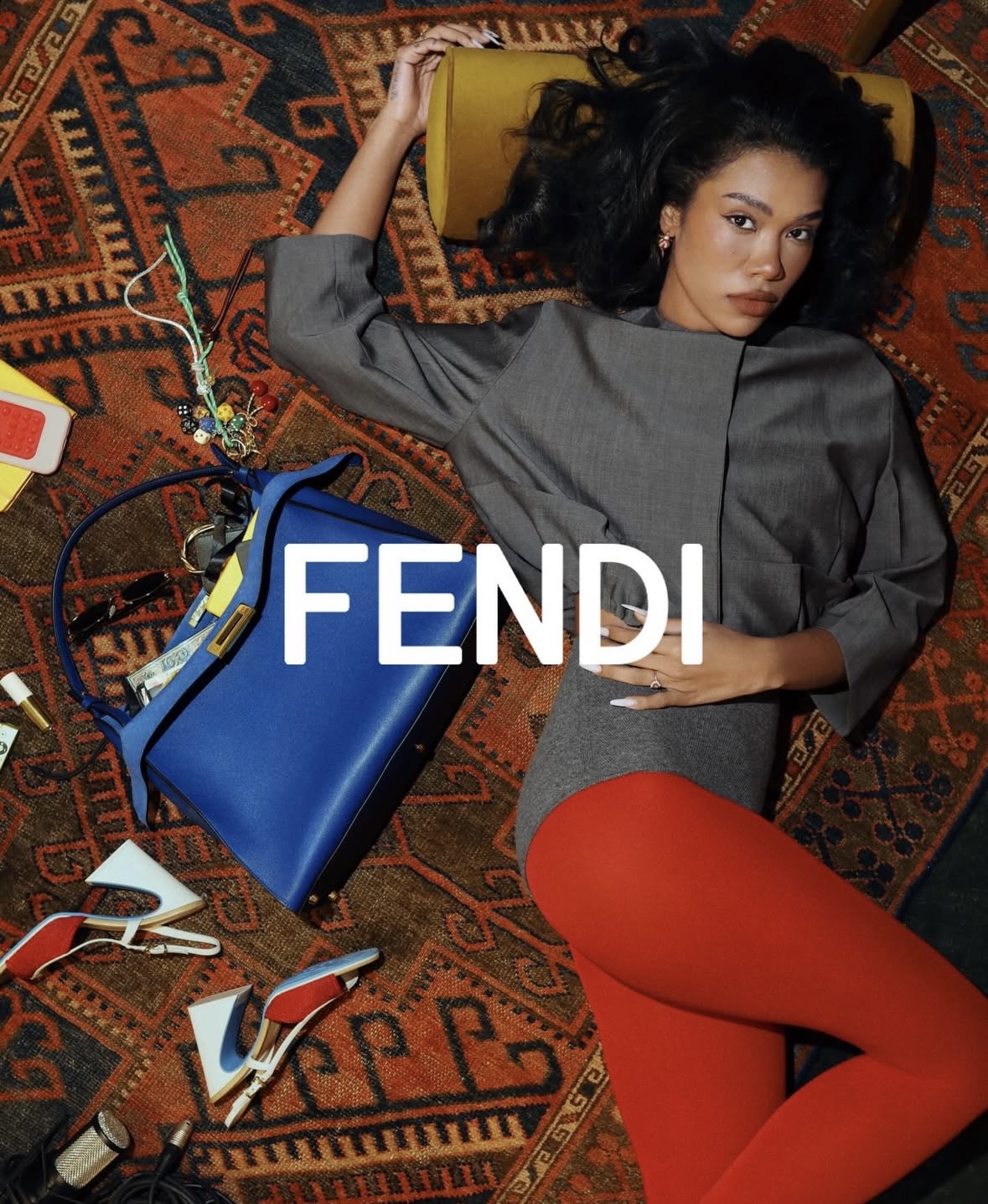 Woman lying on a patterned rug with a blue handbag, electronic devices, and accessories around her.