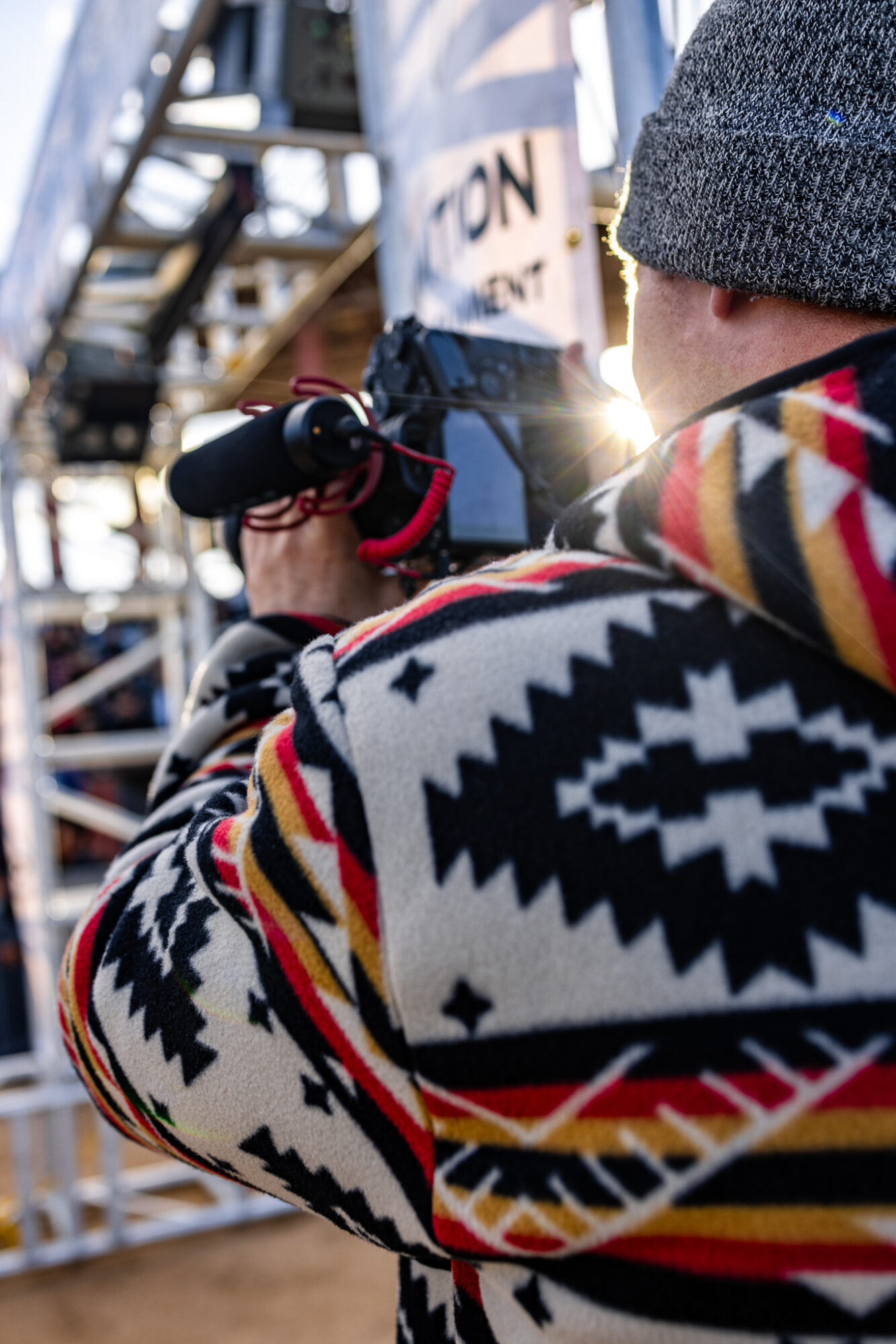 Person wearing a patterned jacket and gray beanie, filming with a camera at an outdoor event with metal structures.