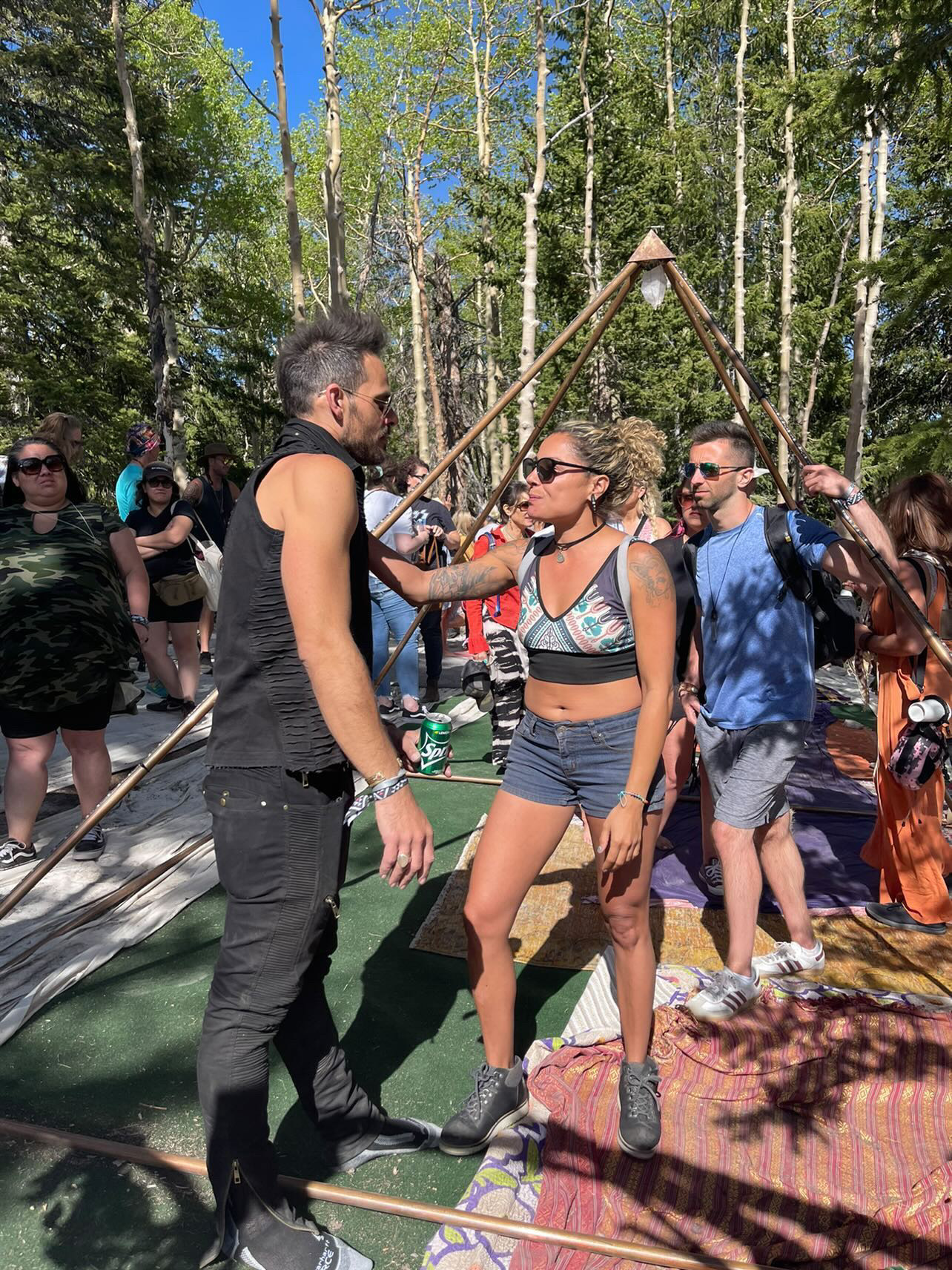 Group of people outdoors in a forested area, some standing and others sitting on mats, with trees and blue sky in background.