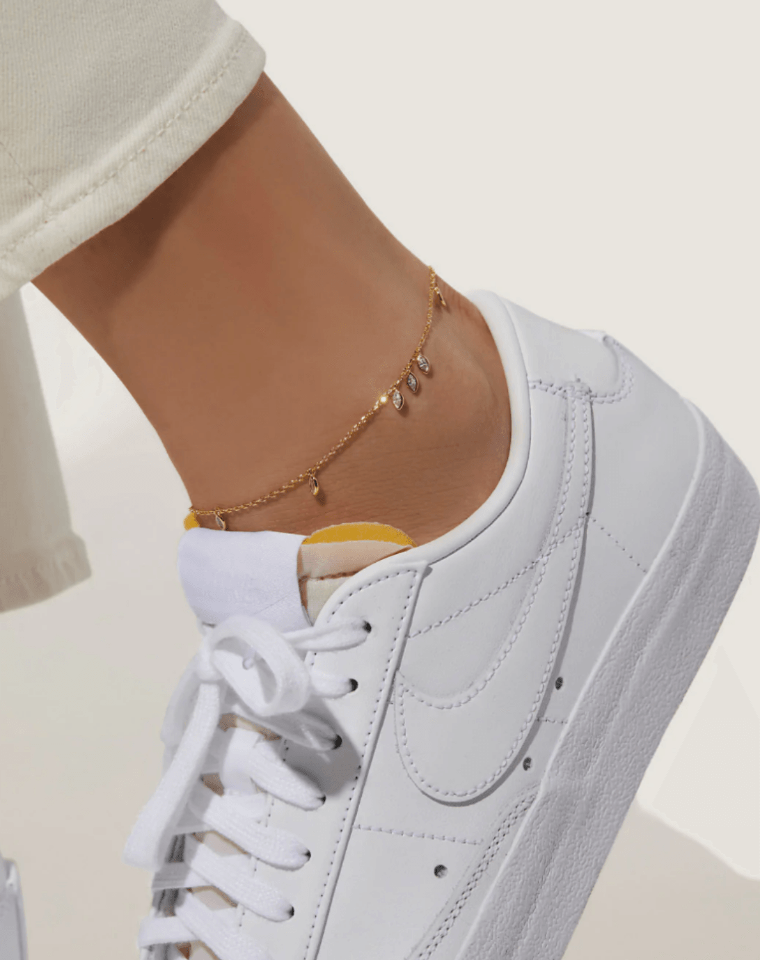 Person wearing a white sneaker and a gold anklet, with beige pants rolled up at the ankle.