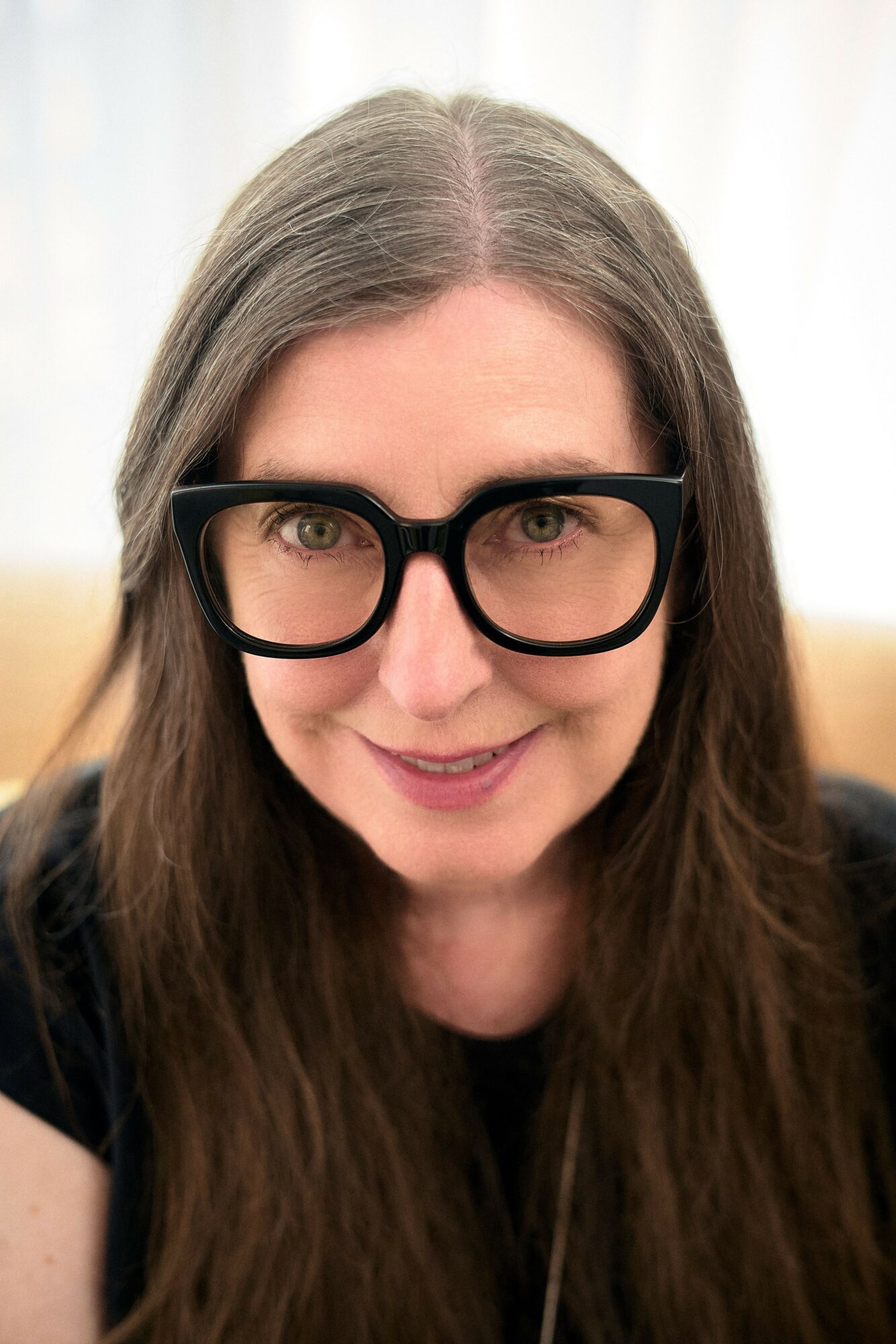 Close-up of a woman with long brown hair wearing black glasses smiling, background is light-colored.