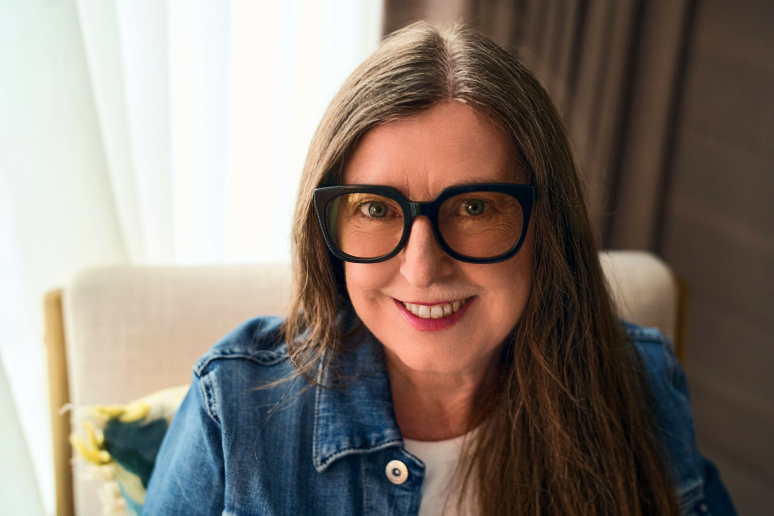Woman with long brown hair, glasses, smiling, wearing a denim jacket, sitting indoors with a light-colored background.