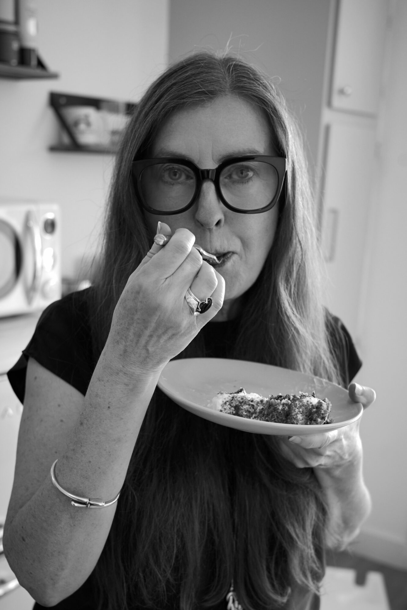 Woman with glasses holding a plate with food, touching her lips, in a kitchen setting, black and white.