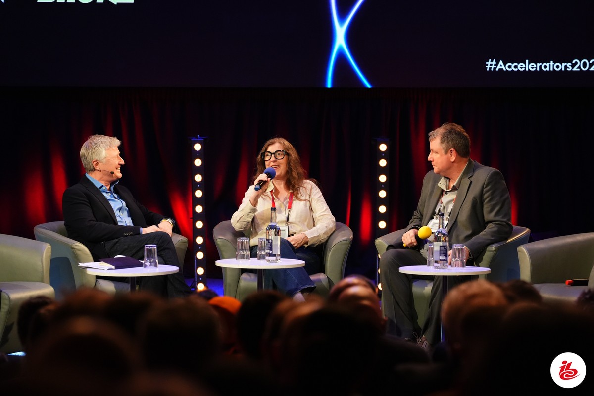 Three people seated on a stage in conversation, with a dark background and audience in foreground.