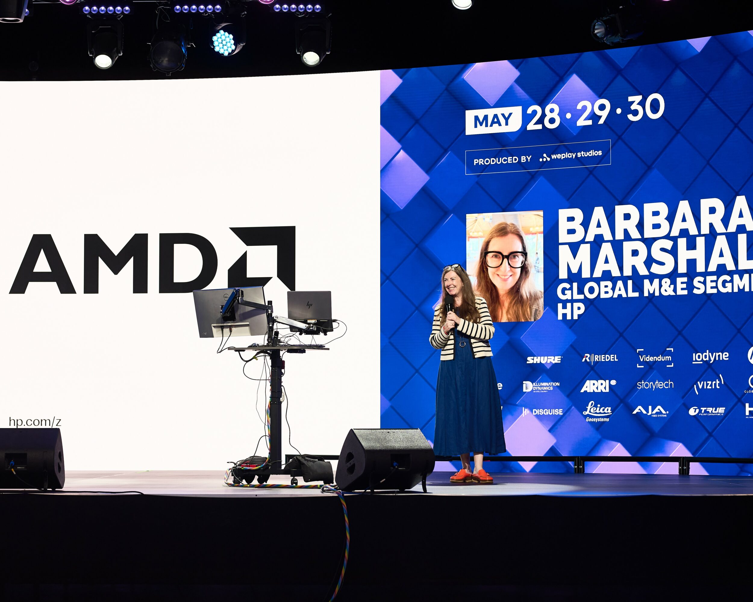 Stage with large screen displaying event dates and speaker's name, woman standing with microphone, AMD logo on screen, stage equipment visible.