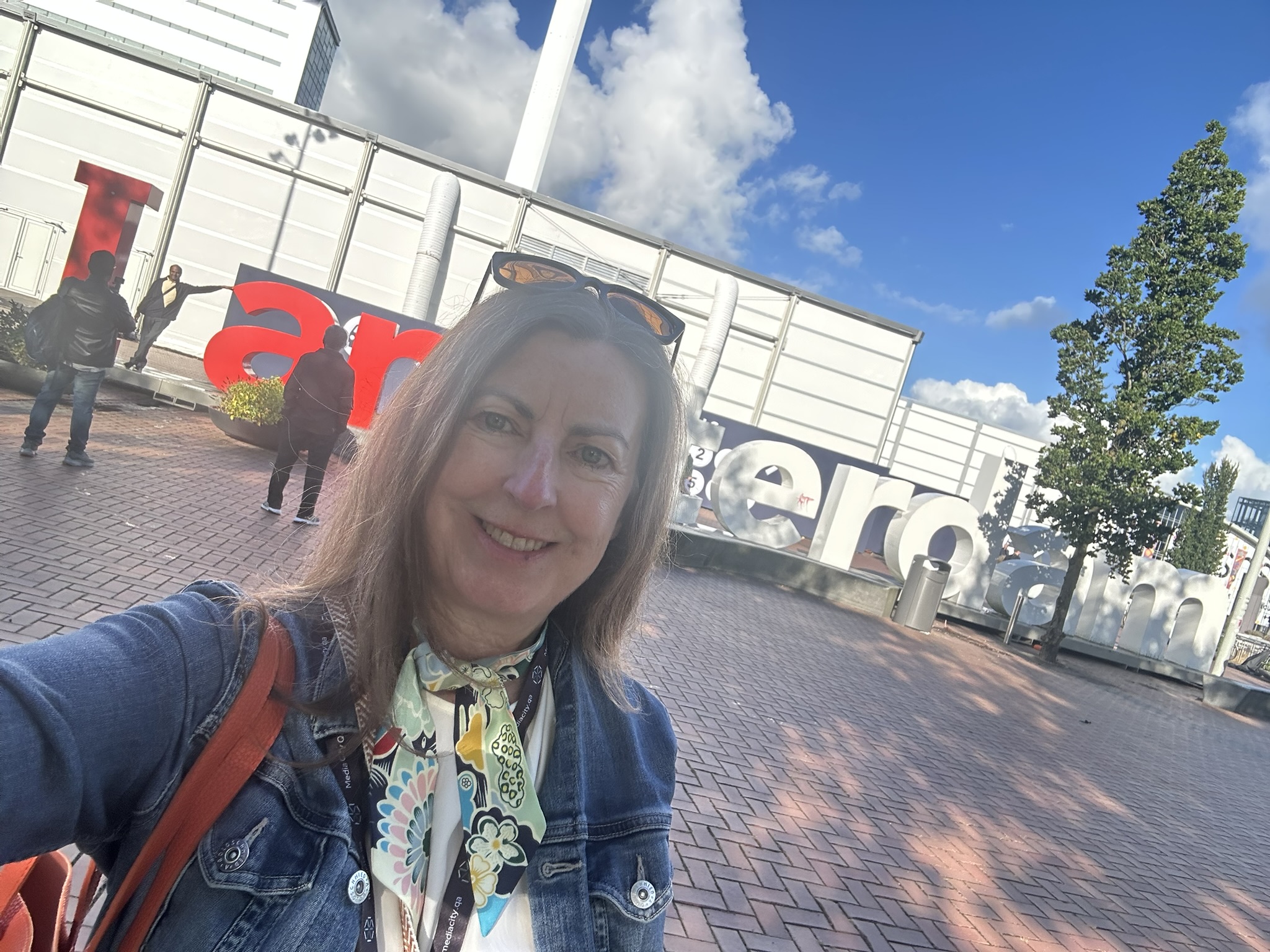 Smiling woman taking selfie outdoors with a large red and white sign in the background, trees, and a blue sky with clouds.